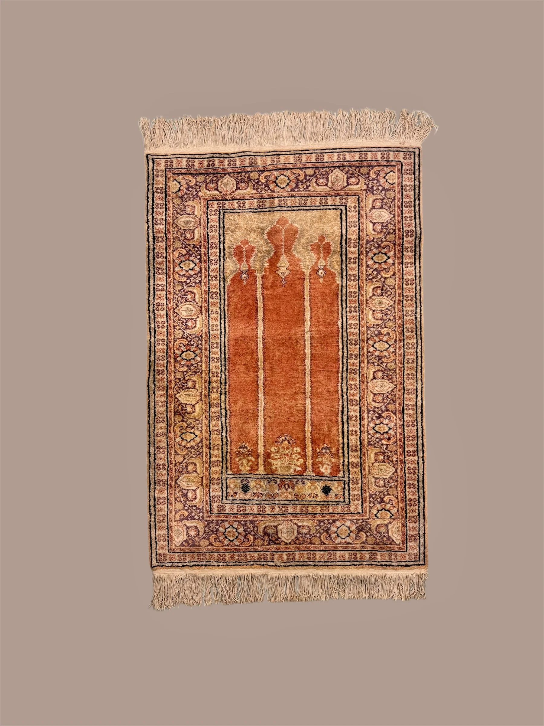 A rectangular area rug with a beige background, featuring an intricate border design with floral patterns in shades of purple, pink, and cream. The central field of the rug has three stylized, symmetrical floral motifs in reddish-orange tones, with s