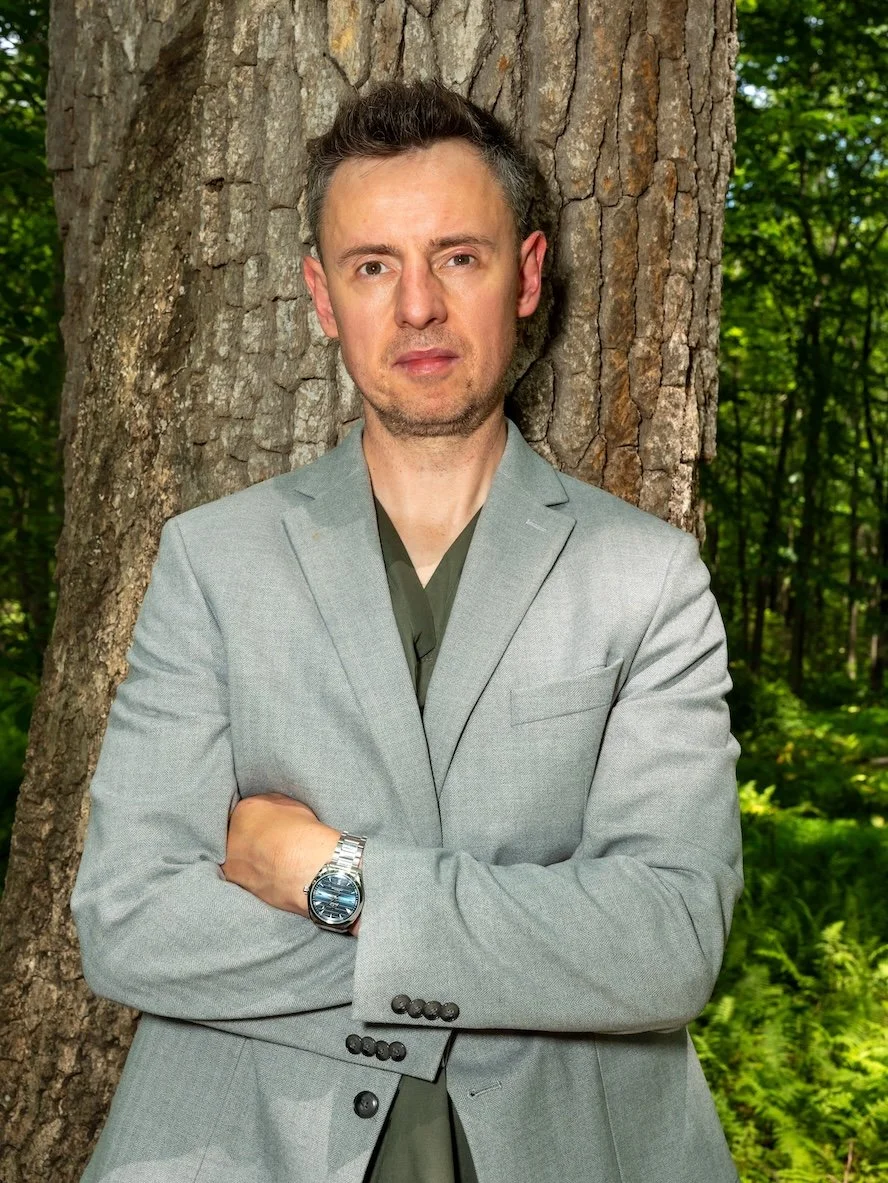A man with short dark hair and a light beard standing outdoors against a large tree trunk, wearing a light gray suit and a dark shirt, with arms crossed and a silver watch on his left wrist.