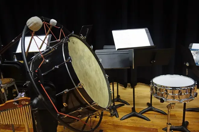 A collection of percussion instruments, including a large bass drum, a snare drum, and a variety of music stands with sheet music, all on a polished wooden stage floor.