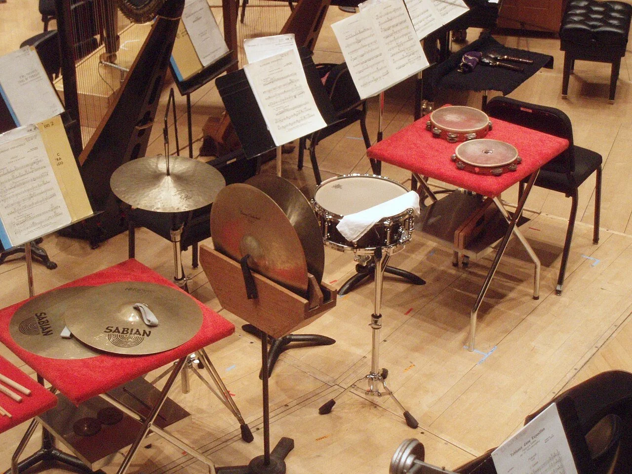 A collection of musical instruments including cymbals, a drum, and tambourines arranged on chairs and stands in a concert hall.