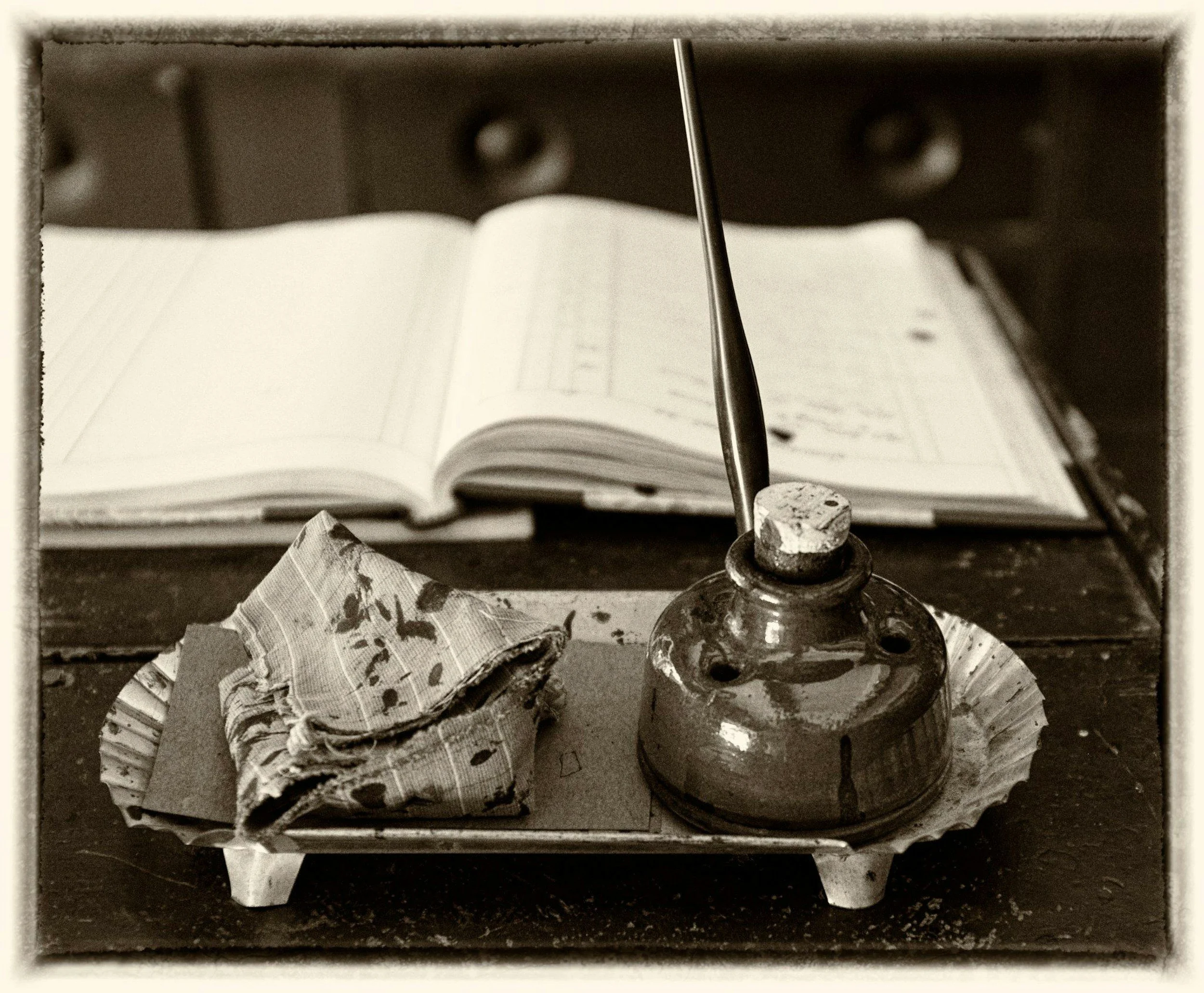 An antique ink well with a pen, next to folded cloth, on a wooden desk with an open ledger in the background.