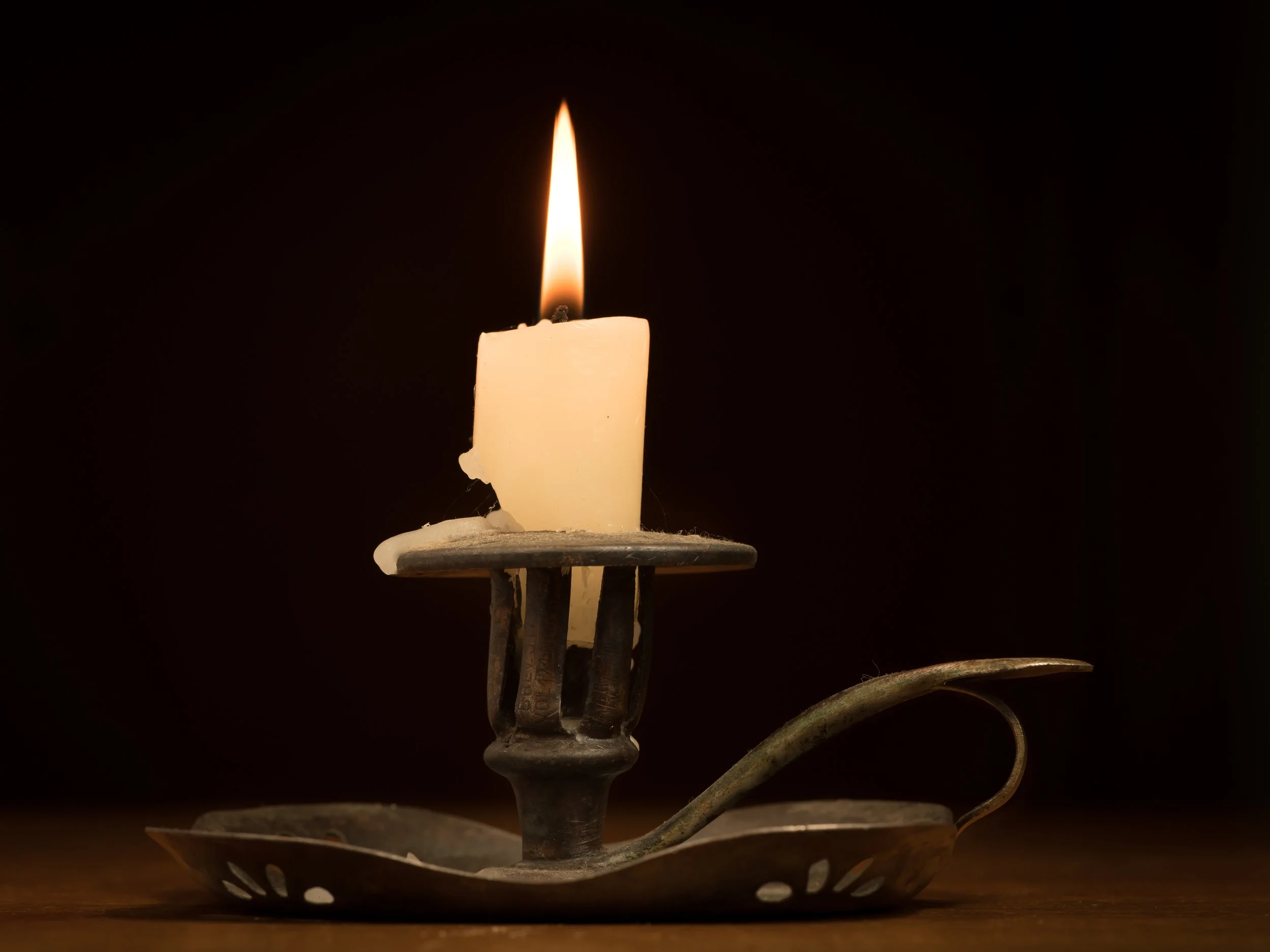 A lit candle on an antique metal candle holder with a leaf design, casting a warm glow against a dark background.