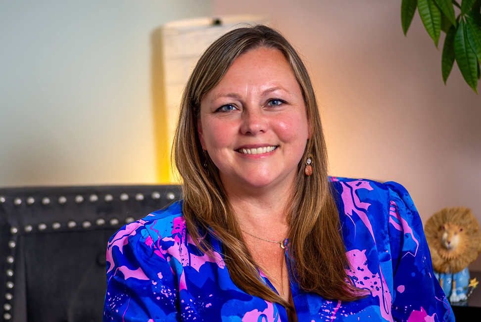 A woman with long light brown hair wearing a blue and purple patterned blouse, smiling in an indoor setting with a plant and a decorative lion in the background.