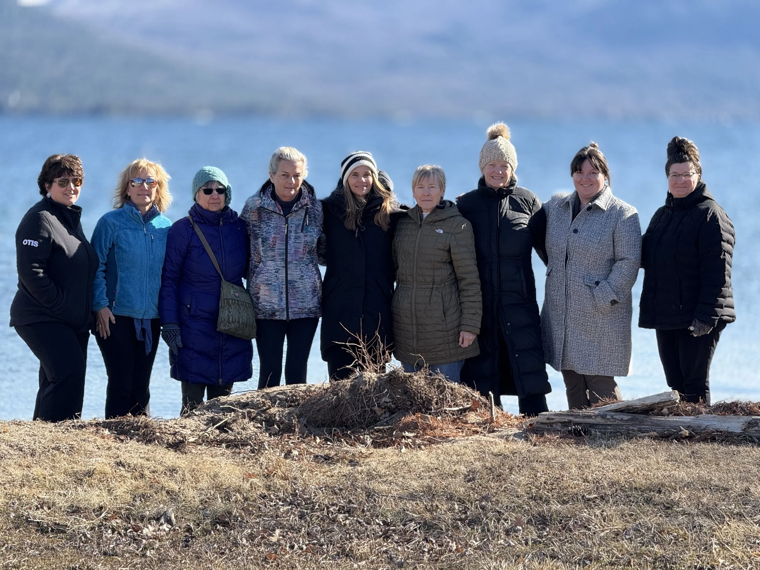 Group of nine women dressed in winter clothing standing outdoors near a body of water with mountains in the background.