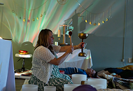 A woman is sitting on a couch in a cozy room, raising a glass in toast with two other glasses on a table, decorated with string lights and balloons.