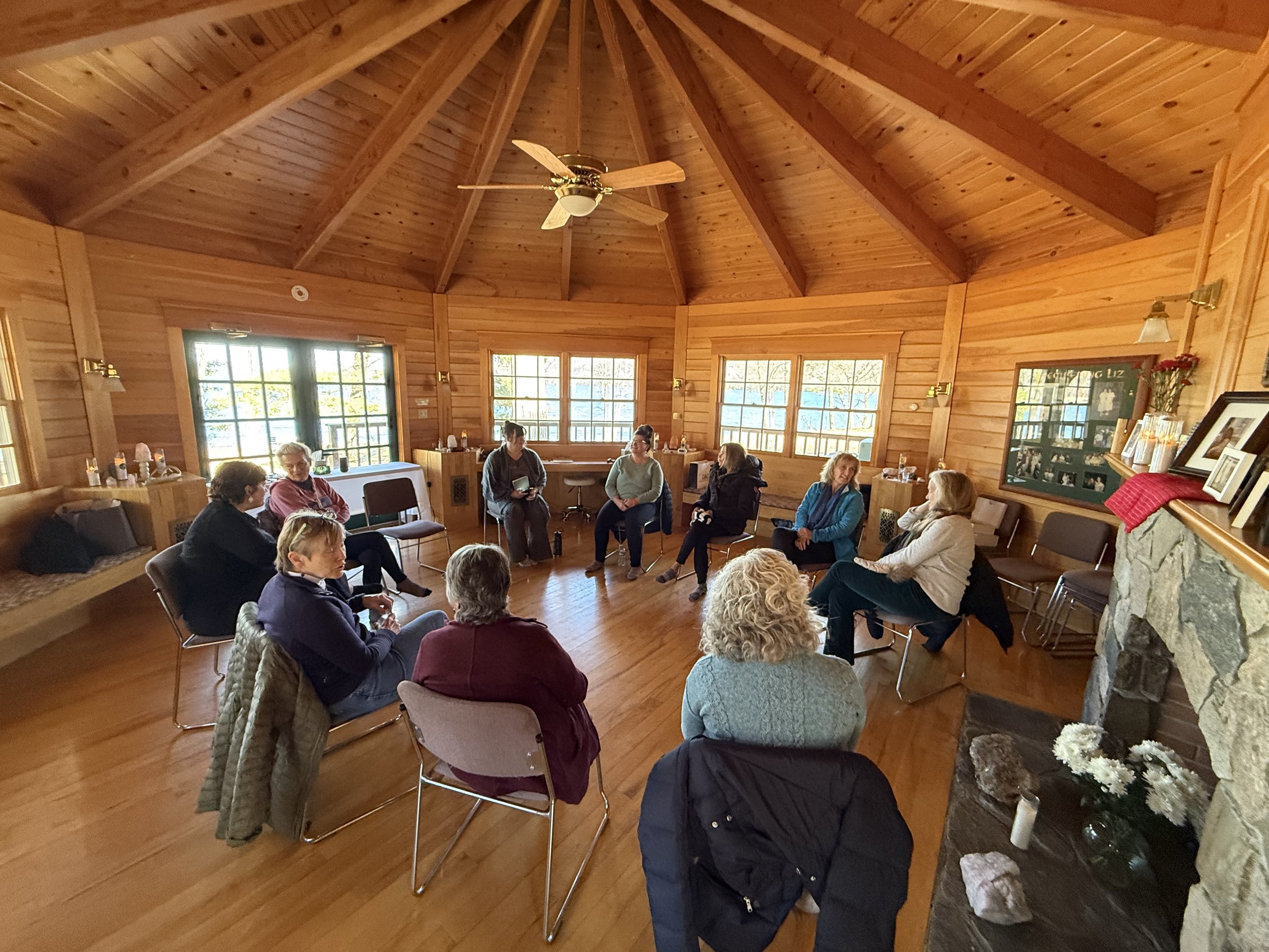 A group of people gathered in a rustic wooden room, sitting in chairs arranged in a semi-circle, participating in a meeting or workshop.