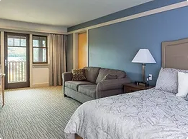Hotel room with a bed, nightstand, lamp, sofa, window, and balcony door with curtains.
