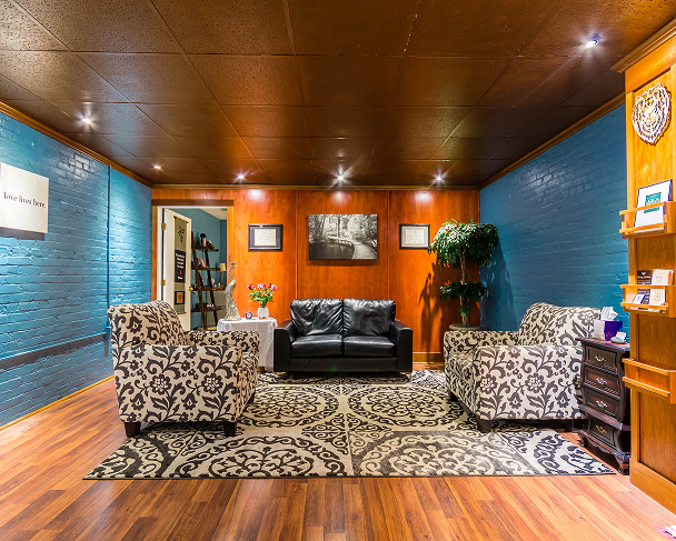 A cozy living room with blue and wood-paneled walls, featuring a black leather sofa, two patterned armchairs, a decorative rug, a potted plant, and framed pictures on the walls.