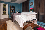A cozy bedroom with a bed covered in white and purple bedding, a small bedside table, a vase of flowers, and a teal accent wall with shelves and decor.