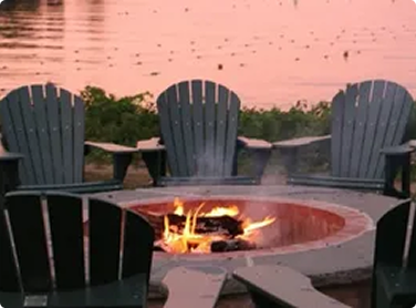 Outdoor fire pit surrounded by Adirondack chairs at sunset with water in the background.