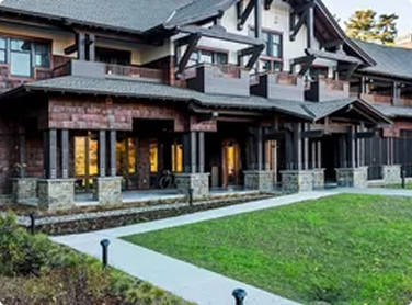 Exterior of a modern multi-story house with stone, wood, and glass elements, surrounded by a manicured lawn and a paved walkway.