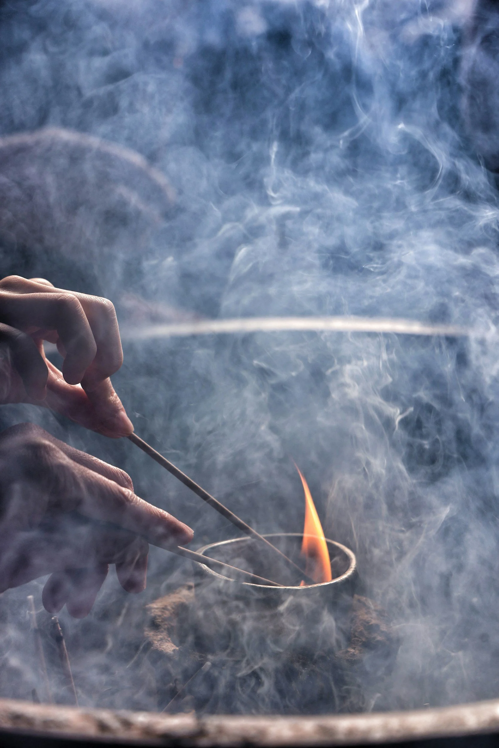Close-up of hands holding a matchstick and a thin metal wire near a small flame, with smoke and steam surrounding the fire