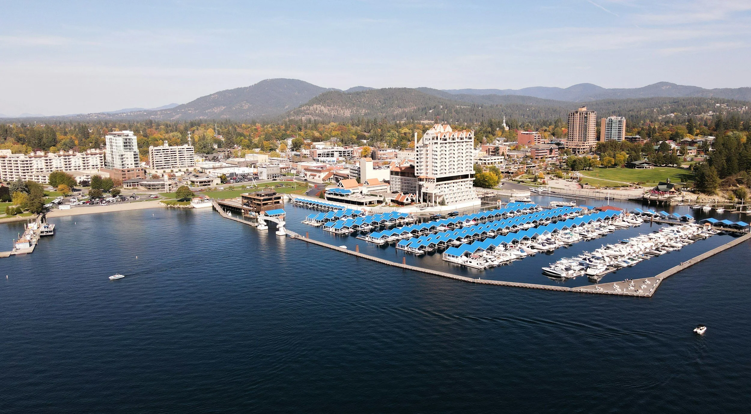 Aerial view of a marina with boats docked under blue canopies, near a city with high-rise buildings, green parks, and mountains in the background.