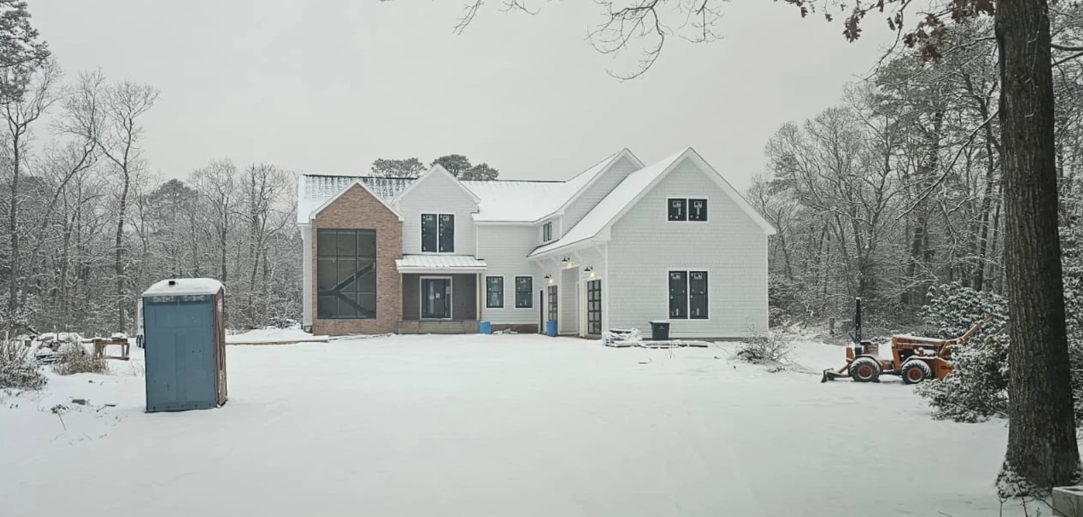 Snow-covered residential house with construction equipment and a portable toilet in the yard, surrounded by leafless trees.