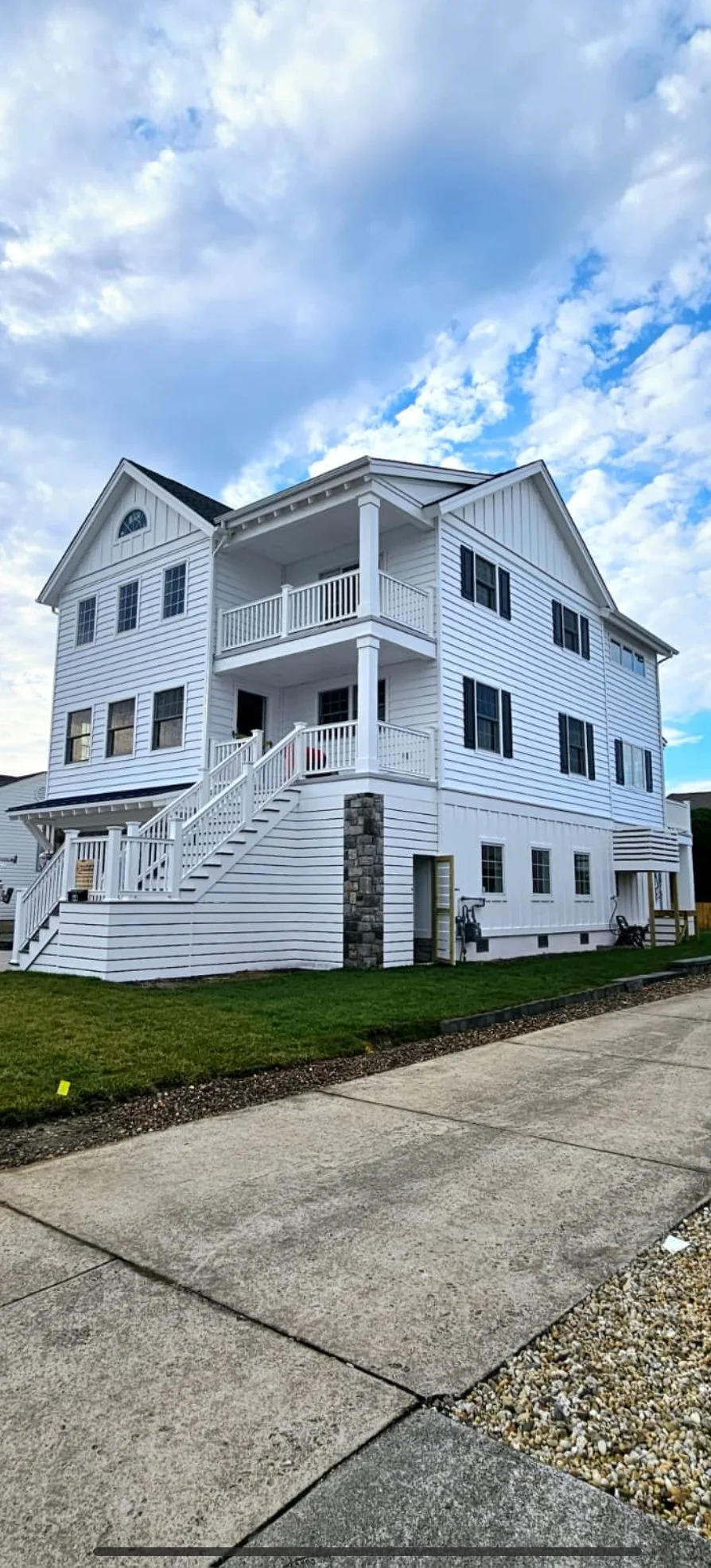 A multi-story white house with black shutters, stairs leading to a front porch, and a partly cloudy sky overhead.