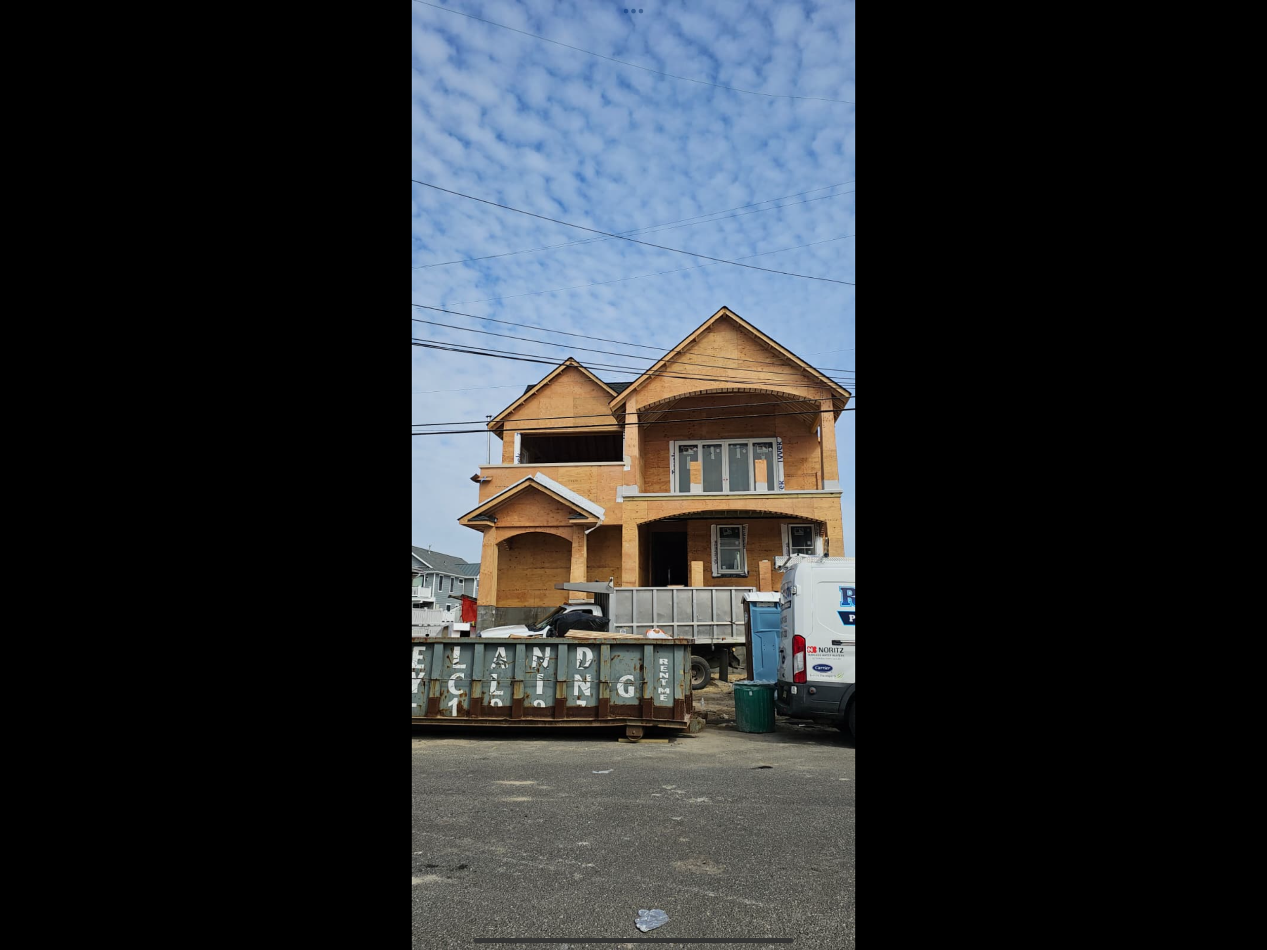 Under construction multi-story residential house with wooden framing, several windows, and a front porch. Construction materials and trucks are in the foreground, with power lines overhead and a partly cloudy sky.