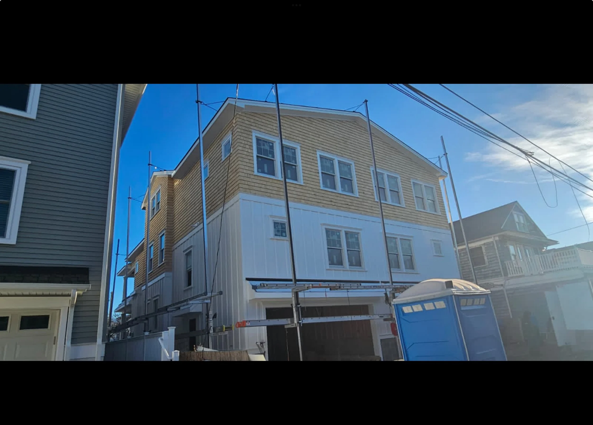 Newly constructed multi-story house with beige and white siding, scaffolding around it, in a neighborhood with other houses and a portable toilet in the foreground.