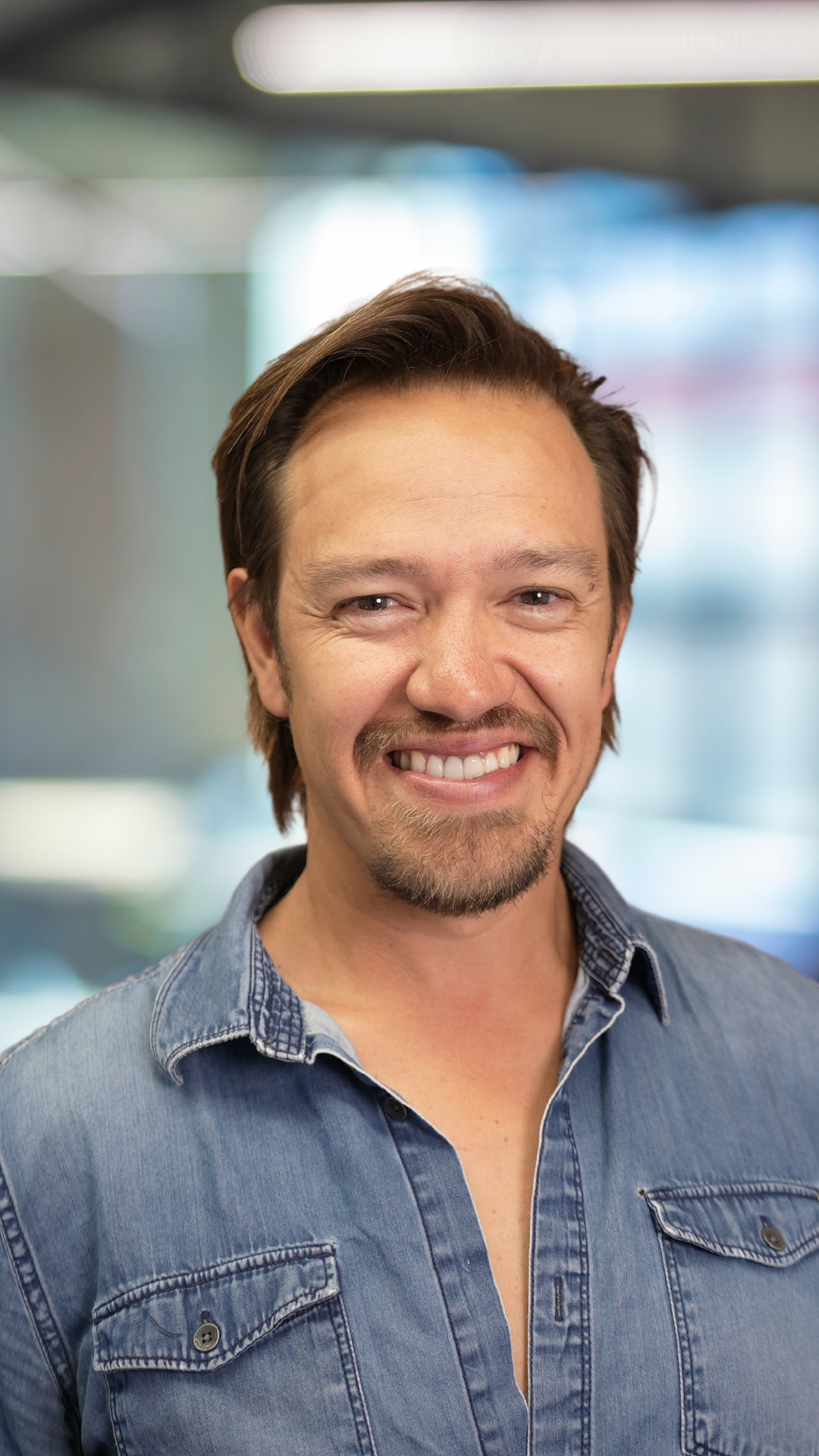A smiling man with brown hair, facial hair, and a goatee, wearing a denim shirt, standing indoors with a blurred background.
