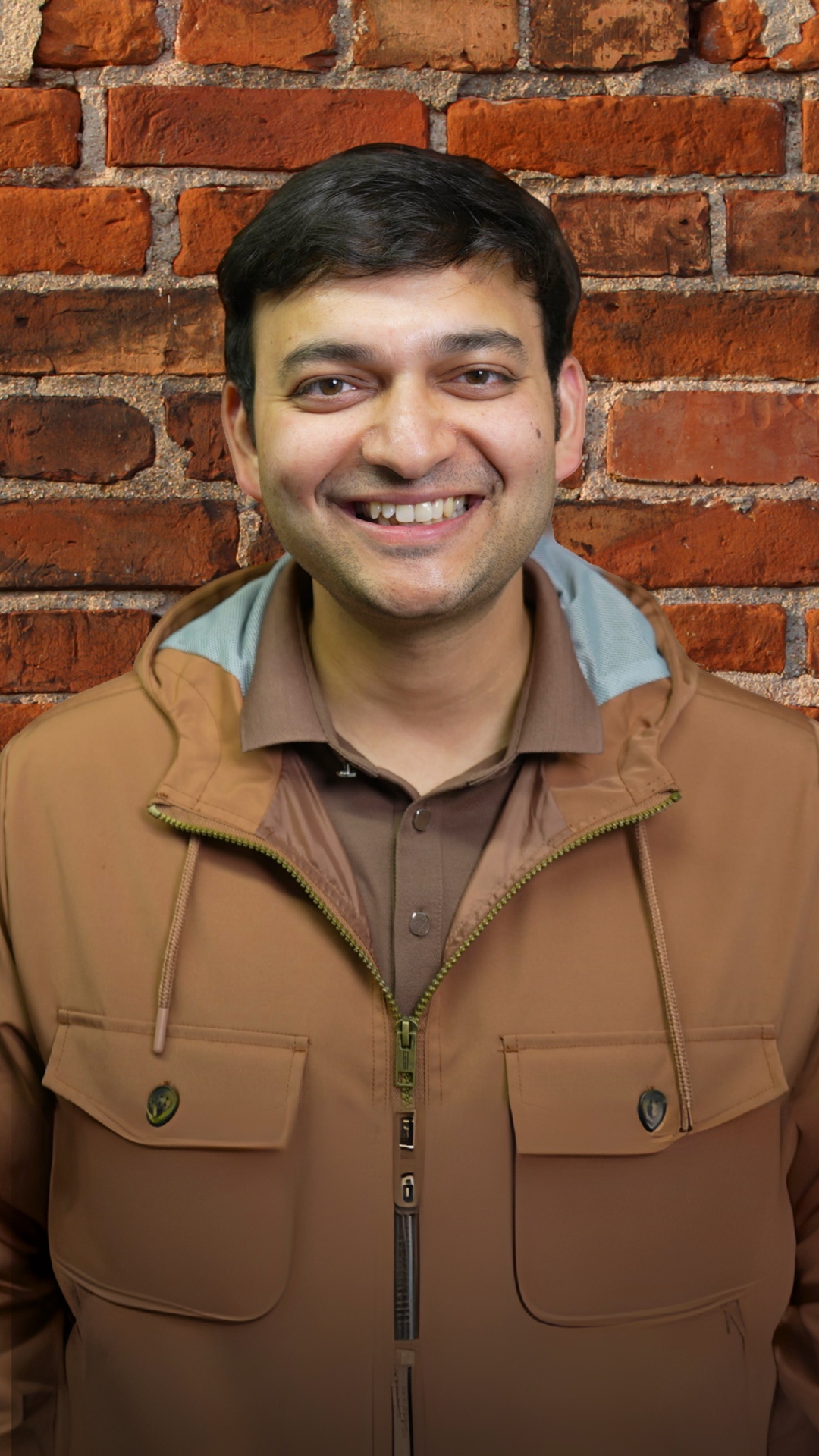 A smiling man with dark hair wearing a brown jacket over a brown shirt, standing in front of a red brick wall.