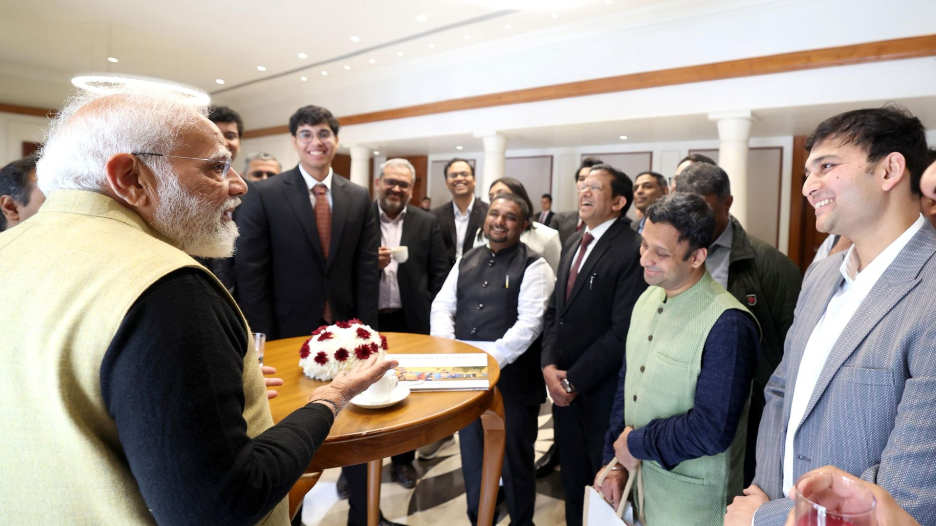 Man in glasses and a navy blue suit being interviewed, sitting at a table with a glass of water, near a window and a tall plant.