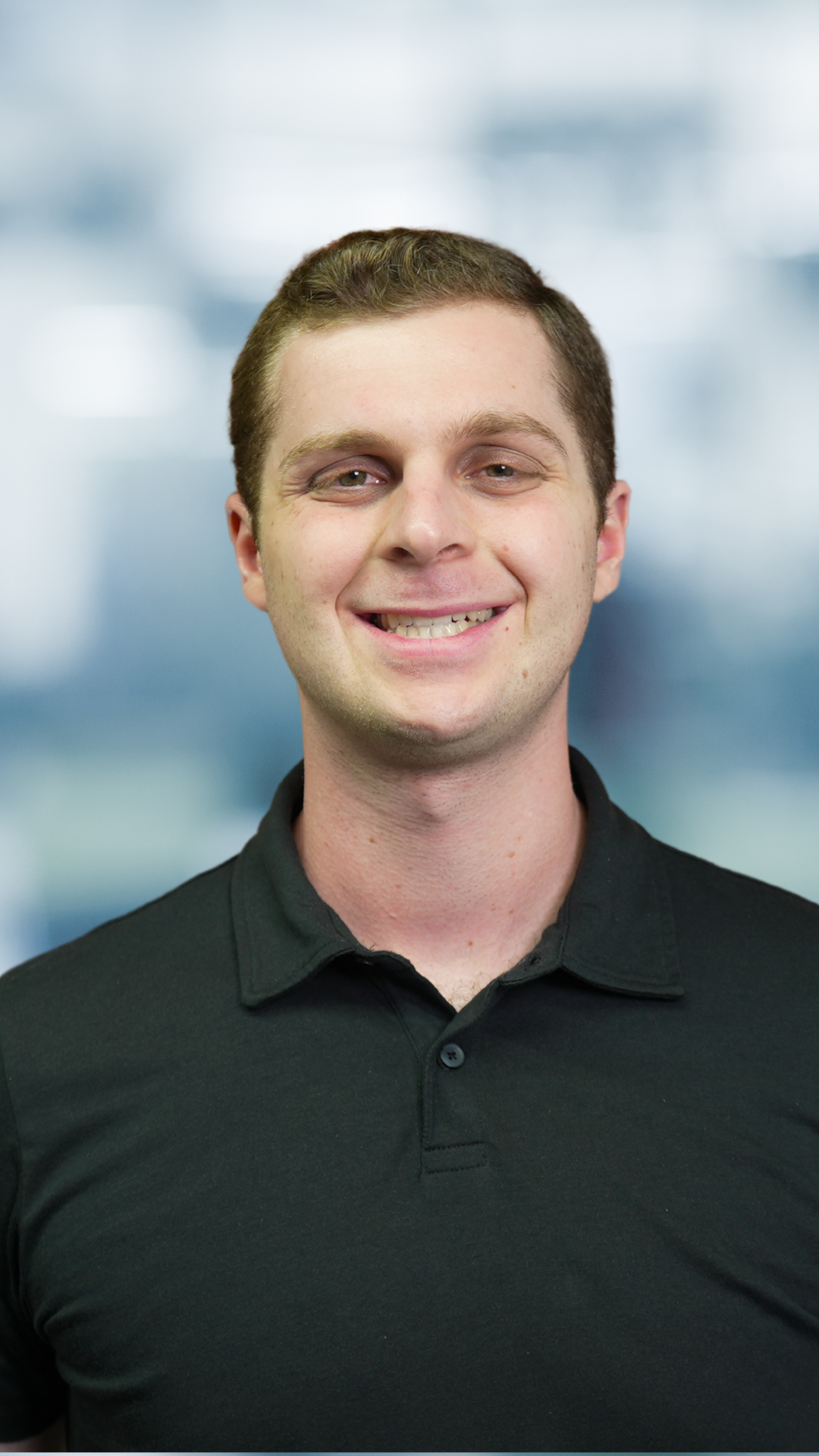 Headshot of smiling young man with short brown hair wearing a black polo shirt, standing against a blurred blue background.