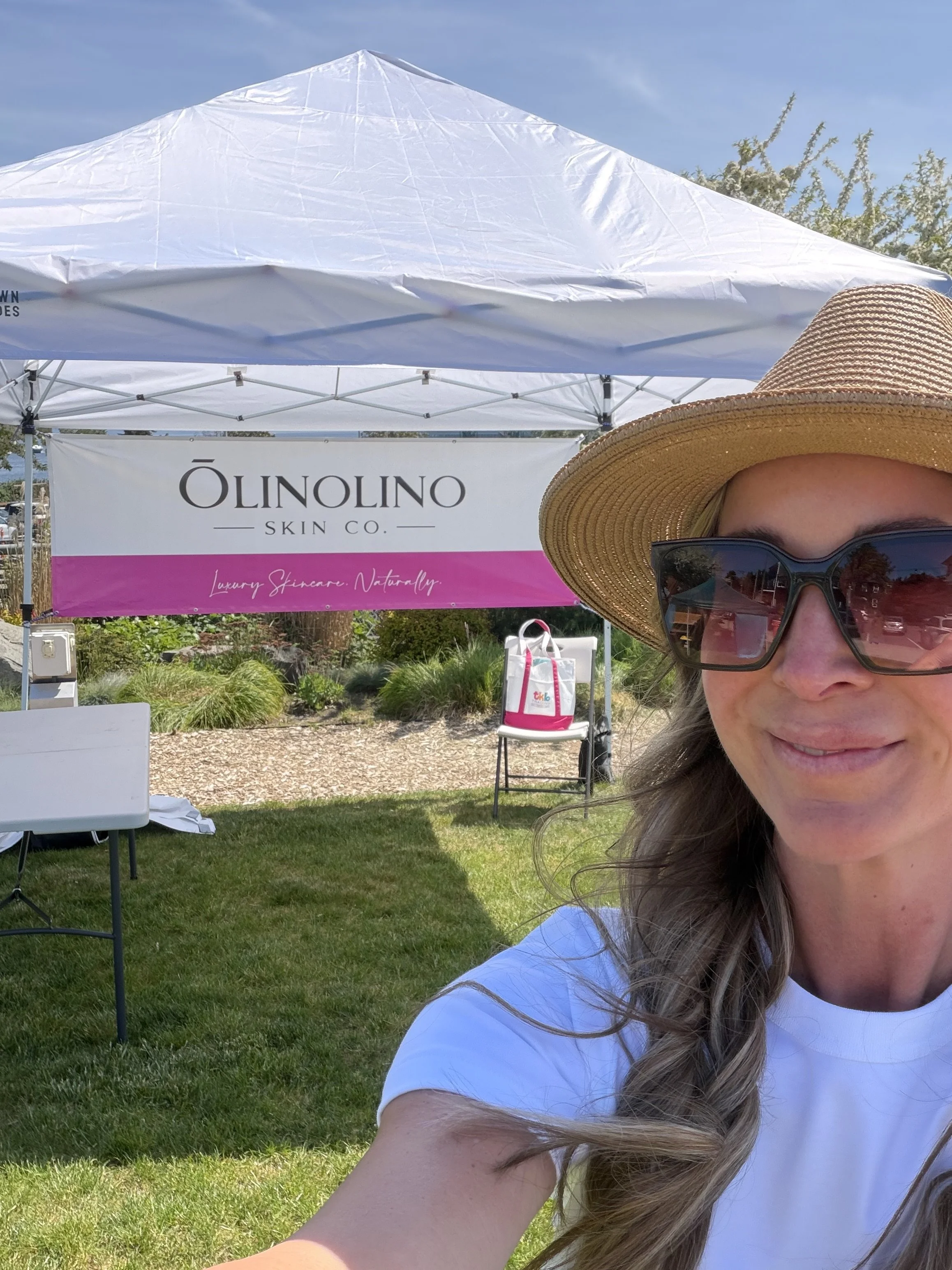 A woman with long light brown hair wearing sunglasses, a straw wide-brim hat, and a white shirt, taking a selfie outdoors under a white canopy tent with a sign for Õlinolino Skin Co., with plants and a sunny sky in the background.