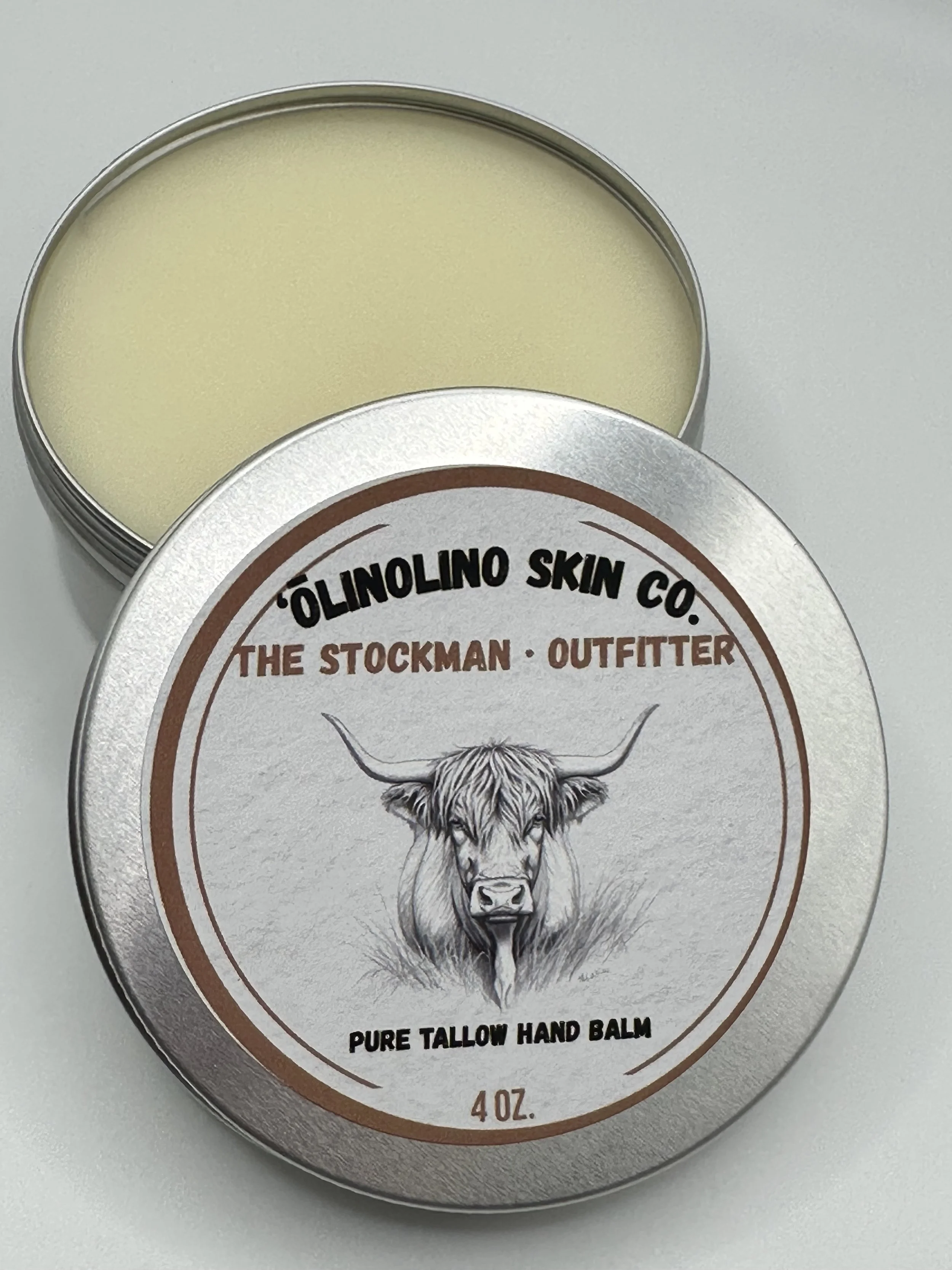 Open tin of hand balm with the lid beside it, labeled 'ÓLÍNOLINO SKIN CO. THE STOCKMAN OUTFITTER PURE TALLOW HAND BALM 4 OZ' and featuring a sketch of a Highland cow.