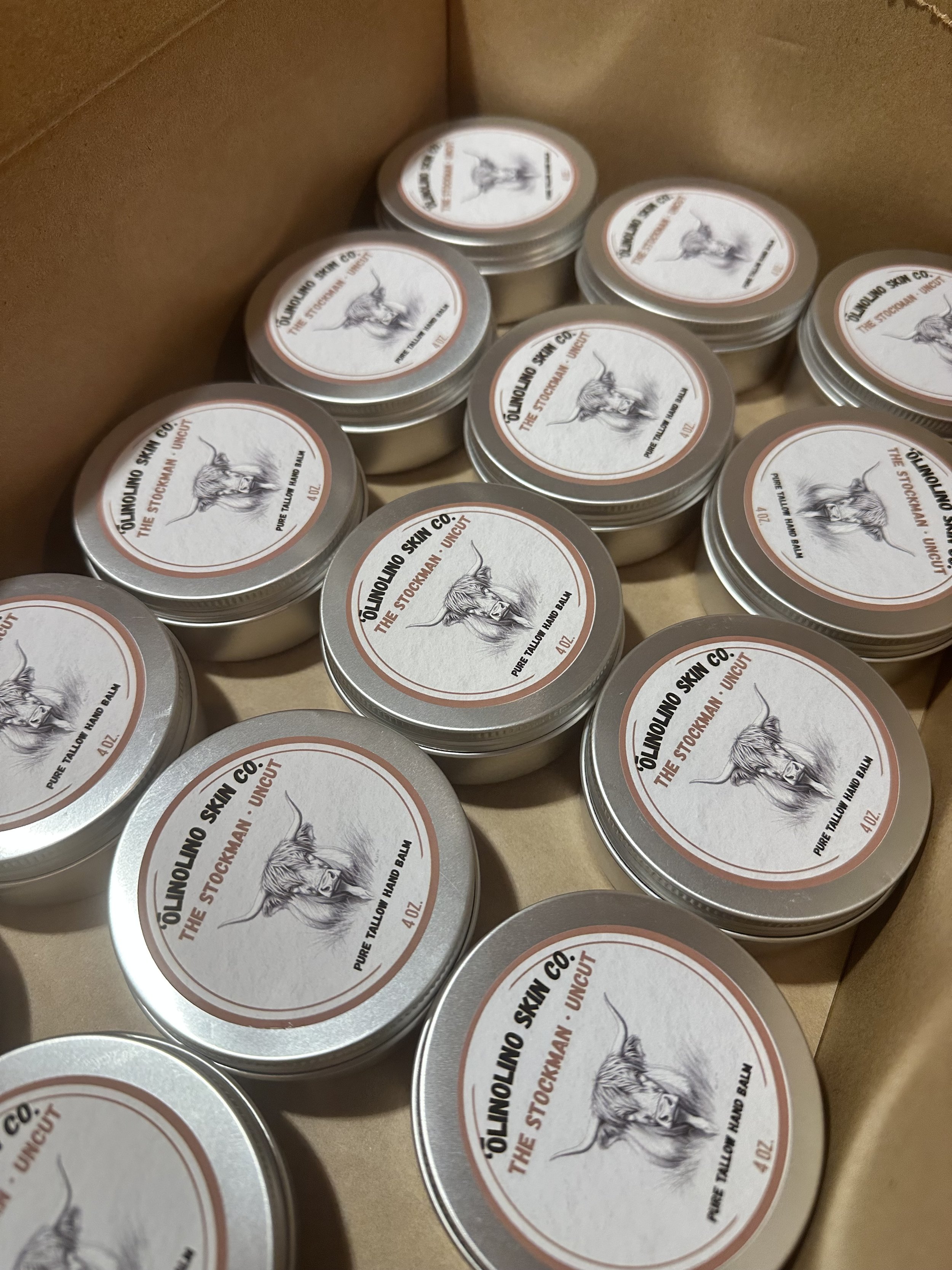 Multiple small tin containers with labels featuring a sketch of a highland cow and text that reads 'ÖLUNLO SKIN CO., THE STOCKMAN UNCUT, PURE TALLOW HAND BALM, 4 OZ' arranged inside a cardboard box.