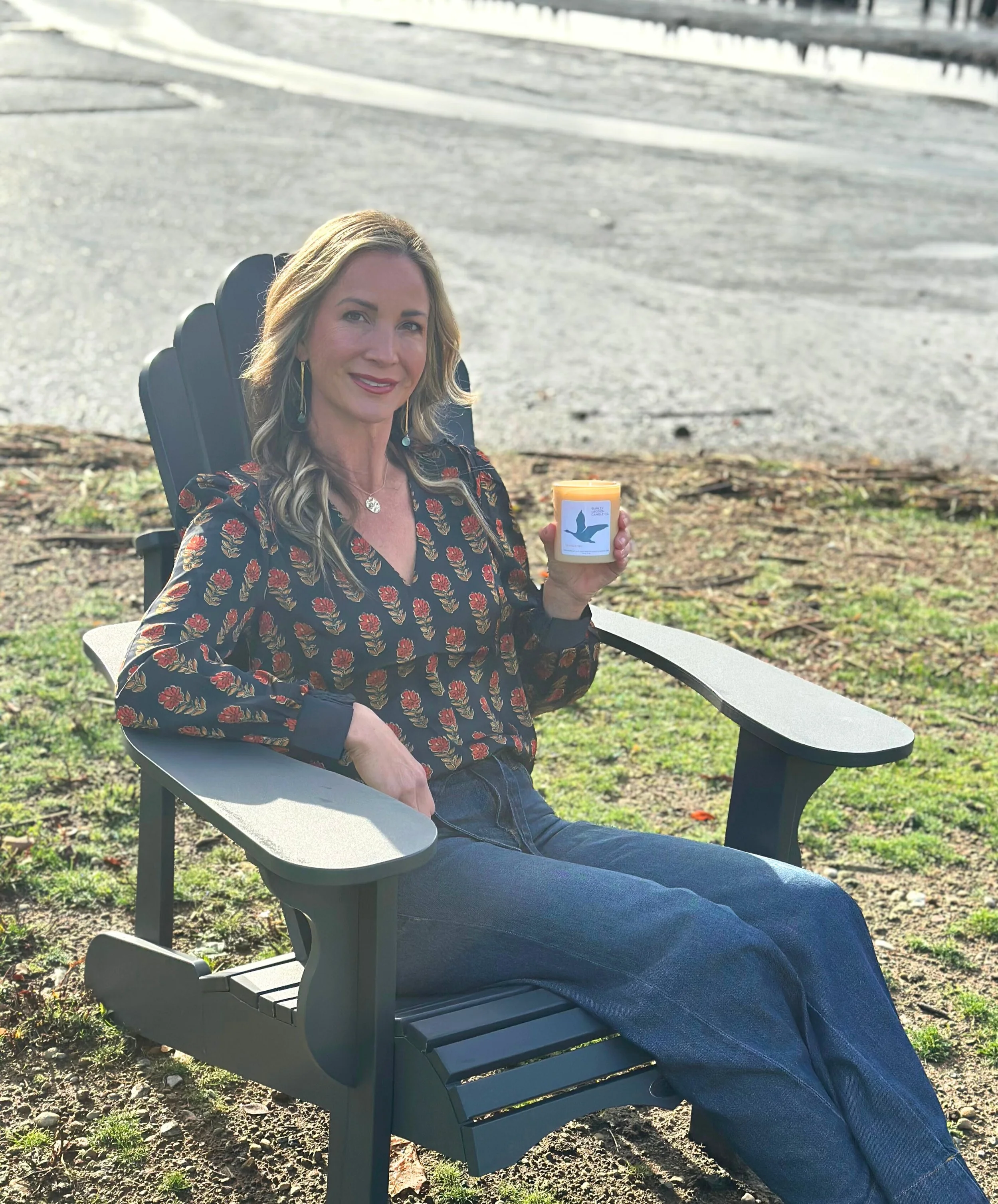 A woman sitting on an outdoor chair near a waterfront, holding a lit candle with a picture of a bird on the label, smiling at the camera.