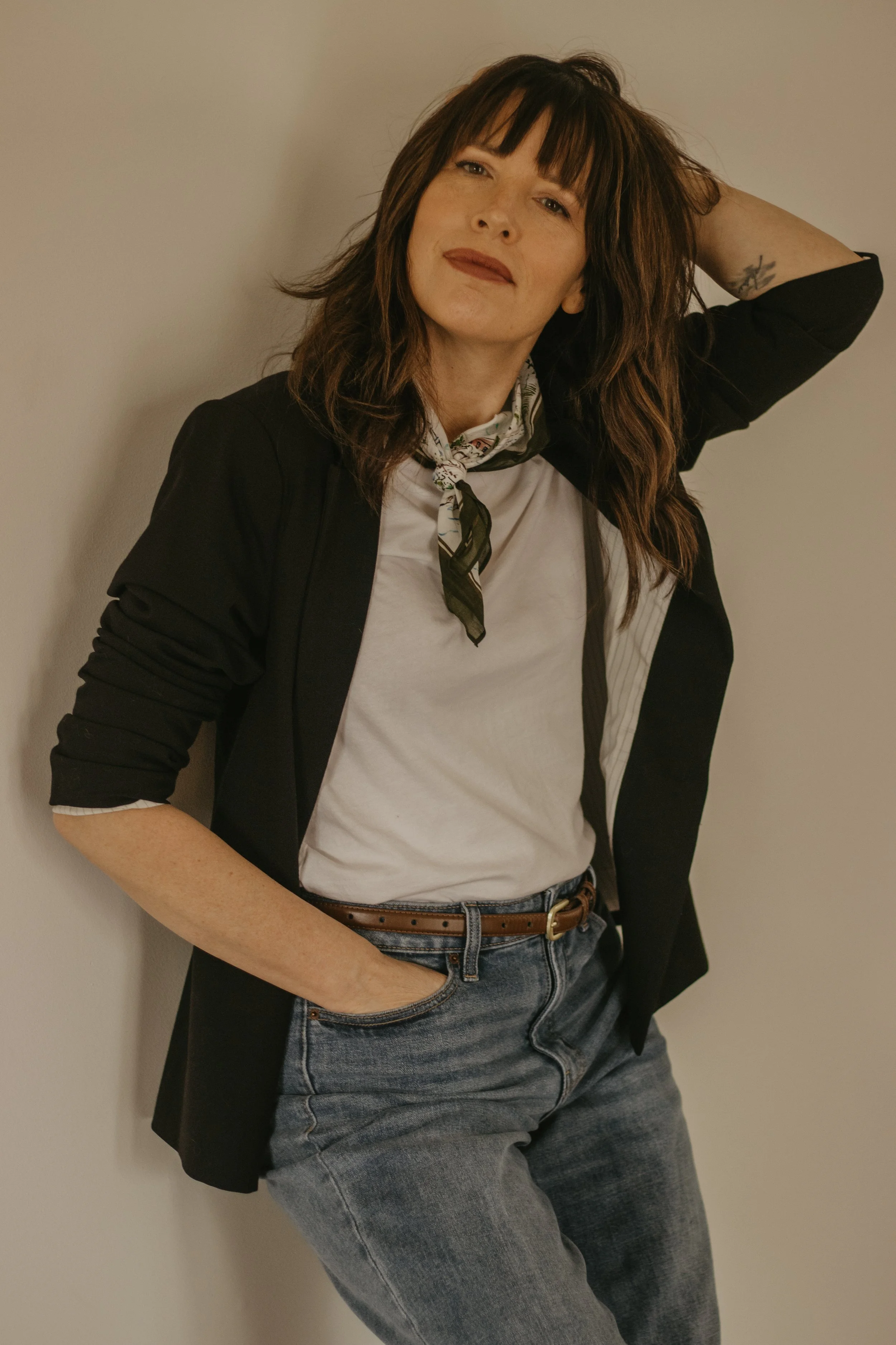 A woman with brown hair and bangs leaning against a wall, smiling slightly with her hand behind her head, wearing a black blazer over a white t-shirt, a patterned scarf, and jeans.