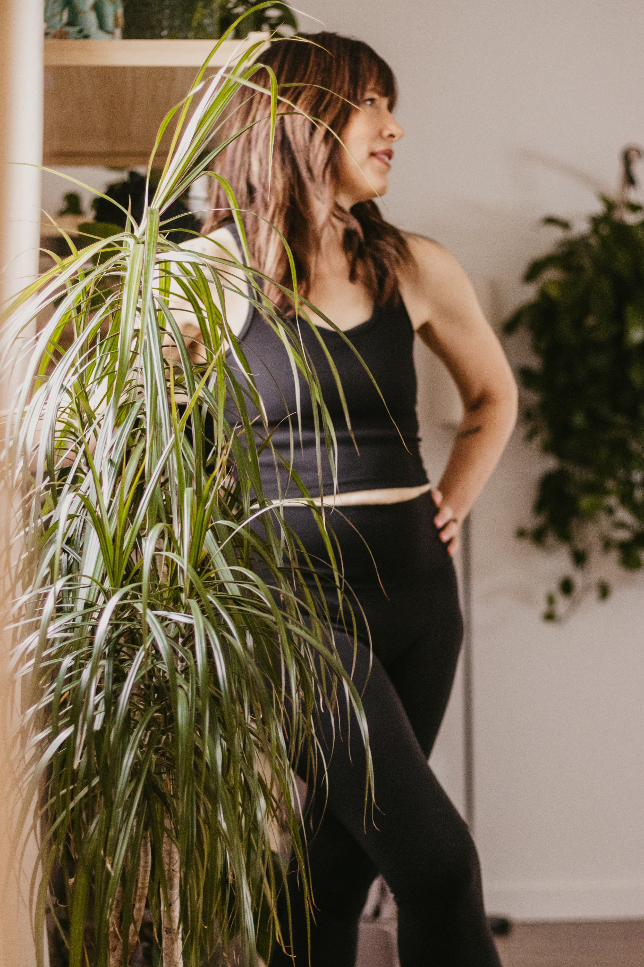 A woman with brown hair and wearing a black sleeveless top standing indoors, with a tall green houseplant in the foreground and other plants in the background.