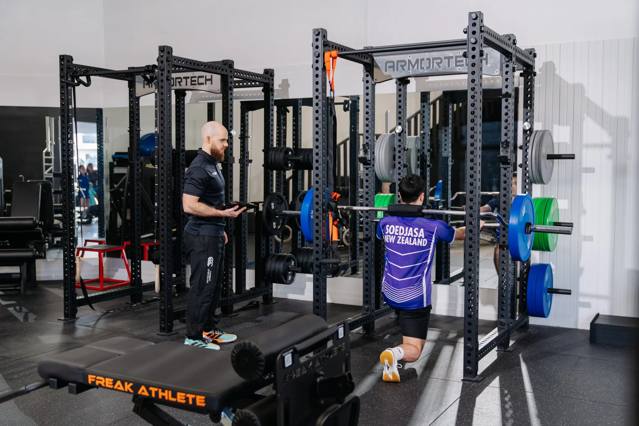 A man doing a kneeling bench press with weights on a bar while being guided by Spencer Precision Fitness personal trainer