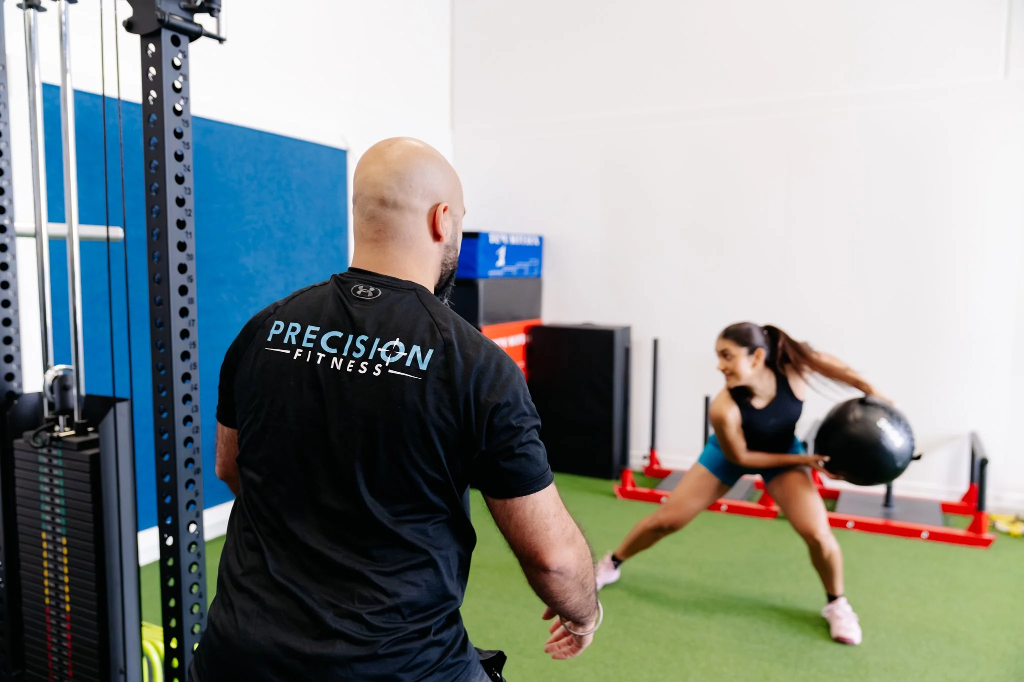A lady throwing a medicine ball against a gym wall while being guided by Precision Fitness personal trainer Coach V