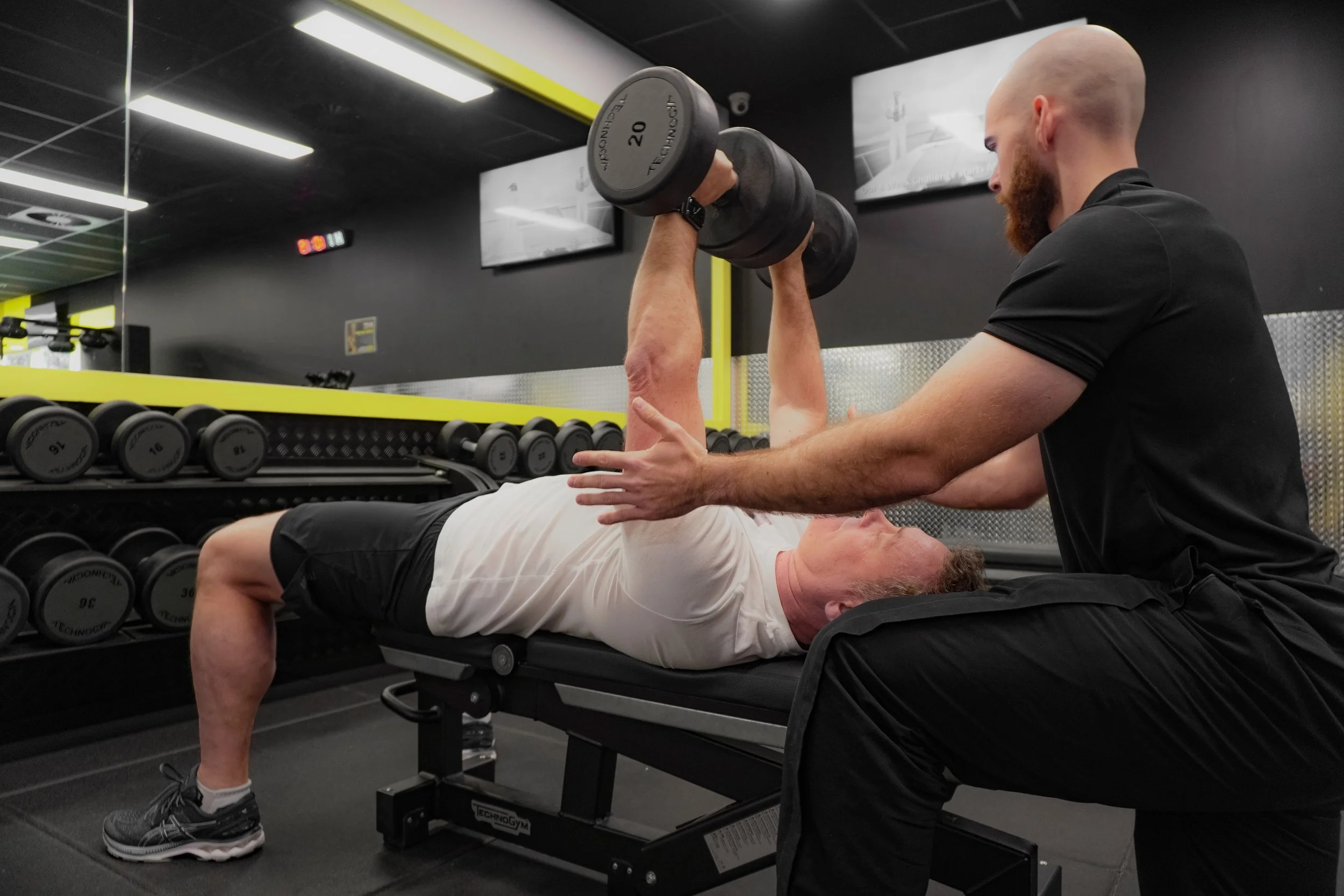 Man on a gym bench lifting weights while be guided by Precision Fitness personal trainer