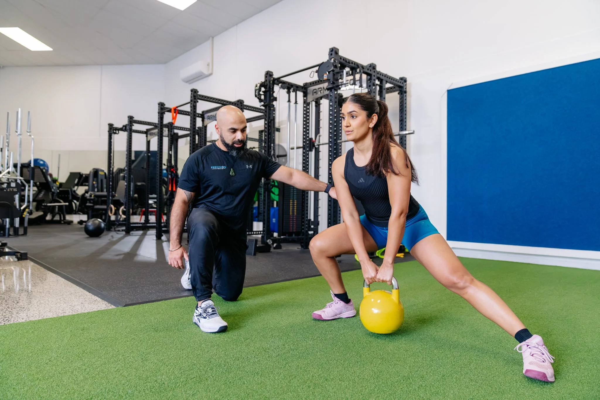 A lady does a split squat with a kettlebell weight while being guided by Coach V Precision Fitness personal trainer