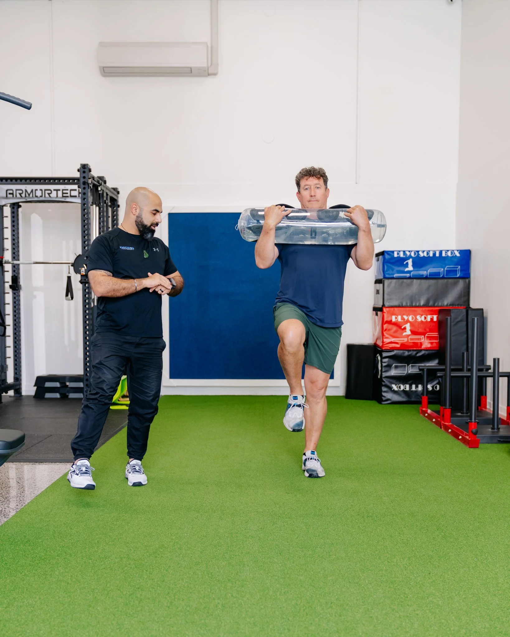 Man carrying a water weight across a gym while being watched by Coach V Precision Fitness personal trainer