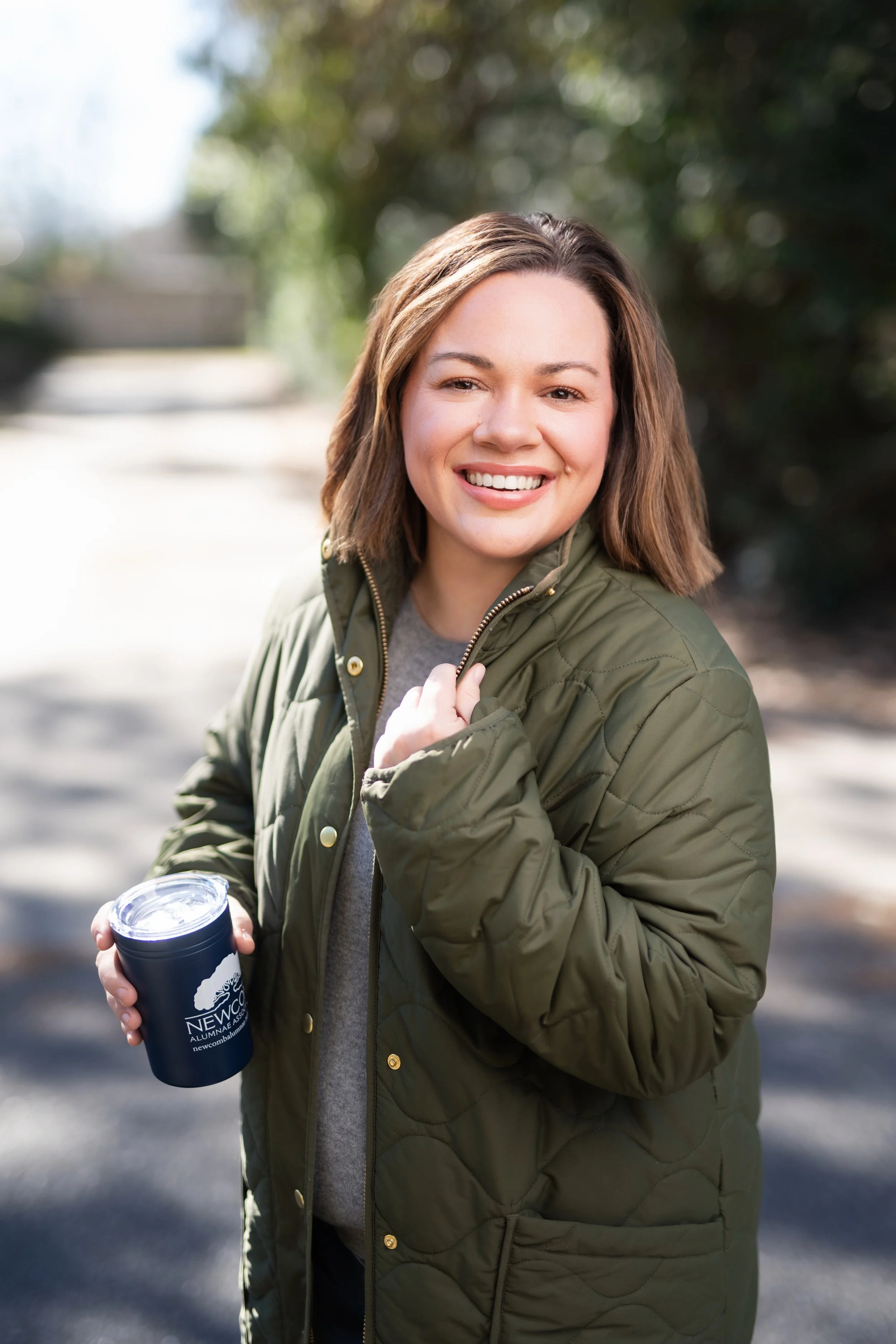 A young woman with brown hair smiling outdoors, holding a black travel mug with a logo, wearing a green quilted jacket on a sunny day.