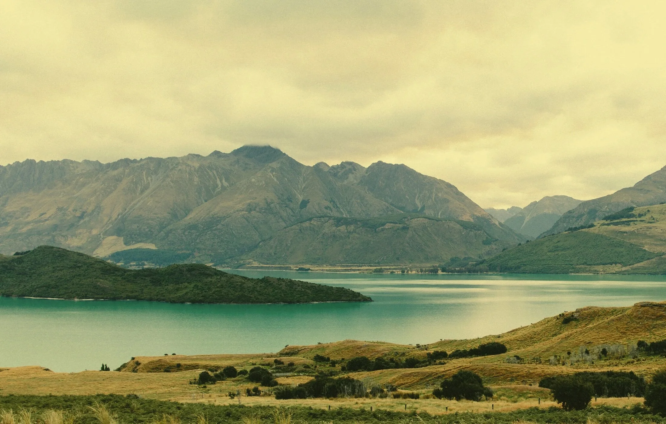 Scenic landscape of a lake surrounded by rolling hills and mountains under cloudy sky
