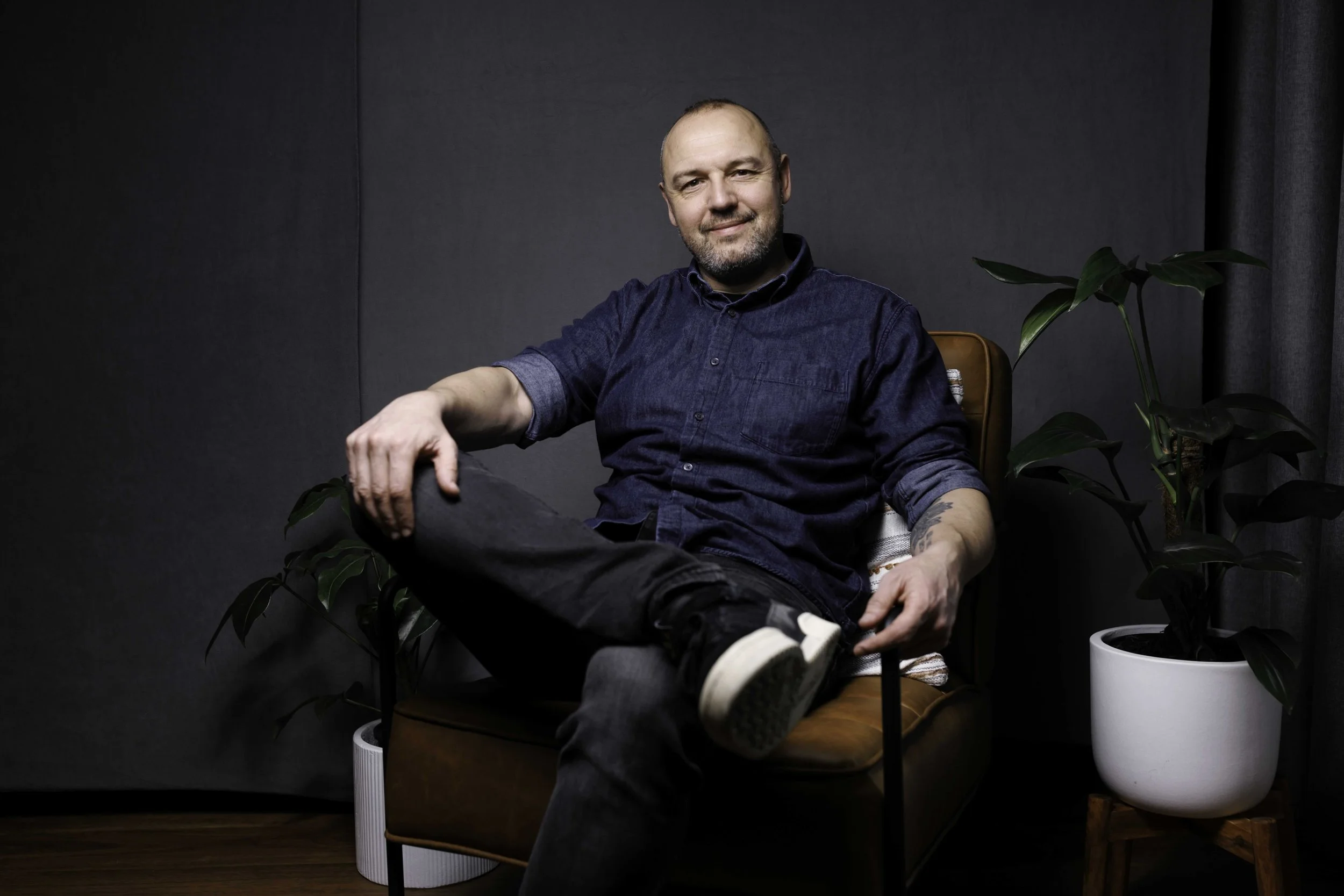 A man sitting in a brown chair with black pants, a dark blue button-up shirt, and white sneakers, smiling with one leg crossed over the other, in a room with a gray wall and dark curtains, flanked by two green potted plants.