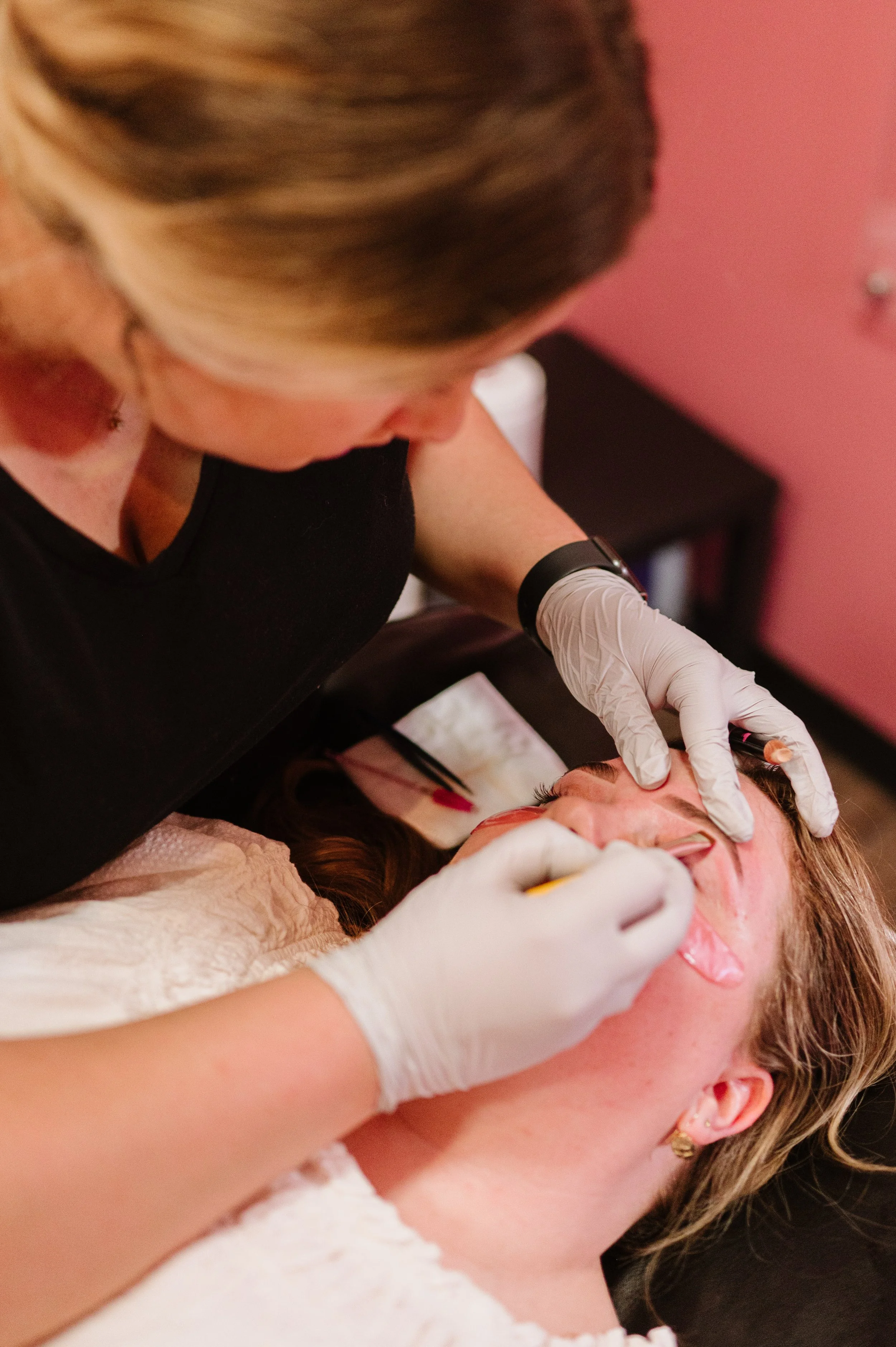 A woman receiving a cosmetic procedure on her lips from a practitioner wearing gloves in a room with pink walls.