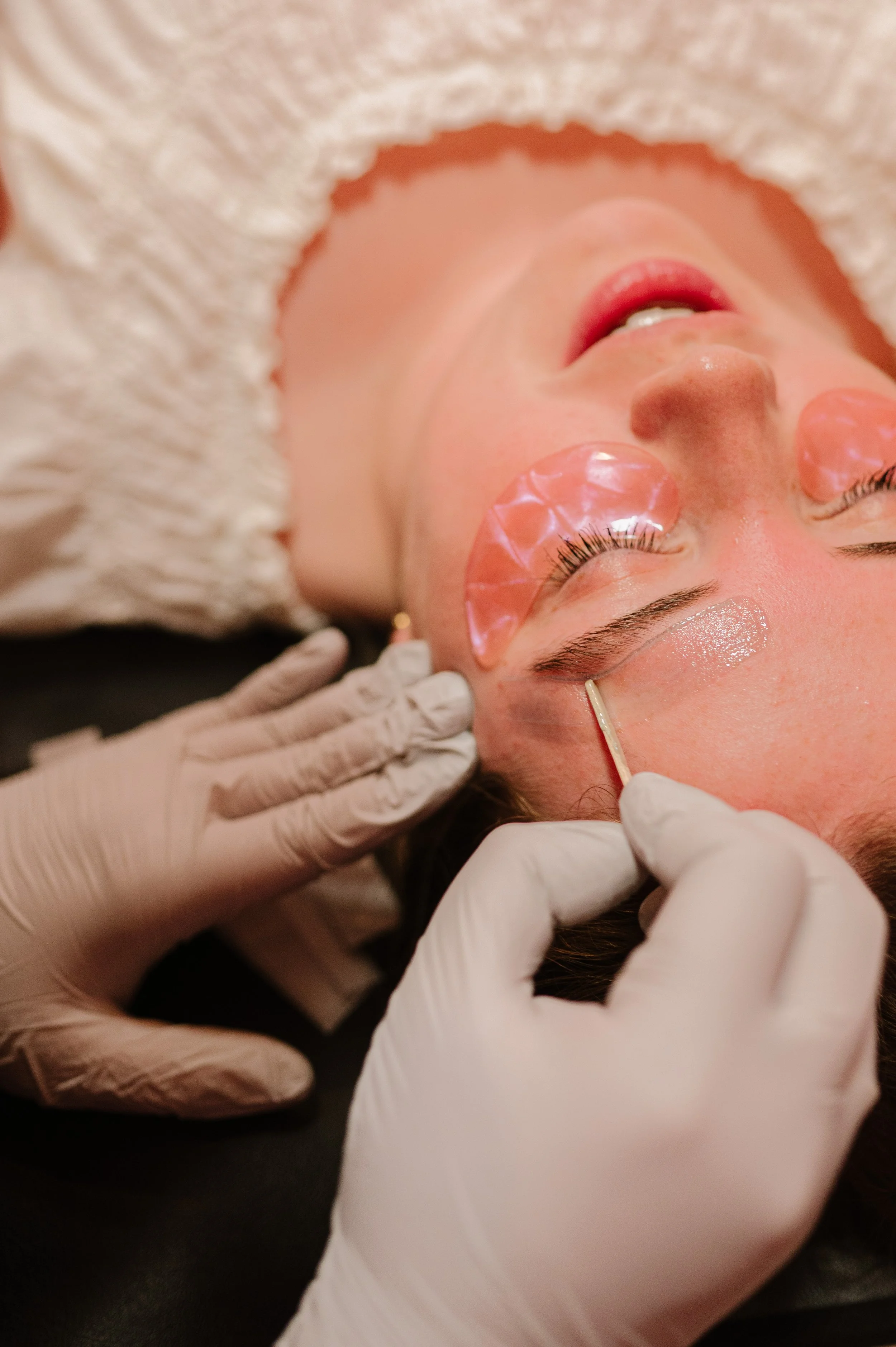 A woman receiving a cosmetic treatment with protective eye patches on her eyelids while a professional applies a product near her eyebrow using a small stick, wearing gloves.