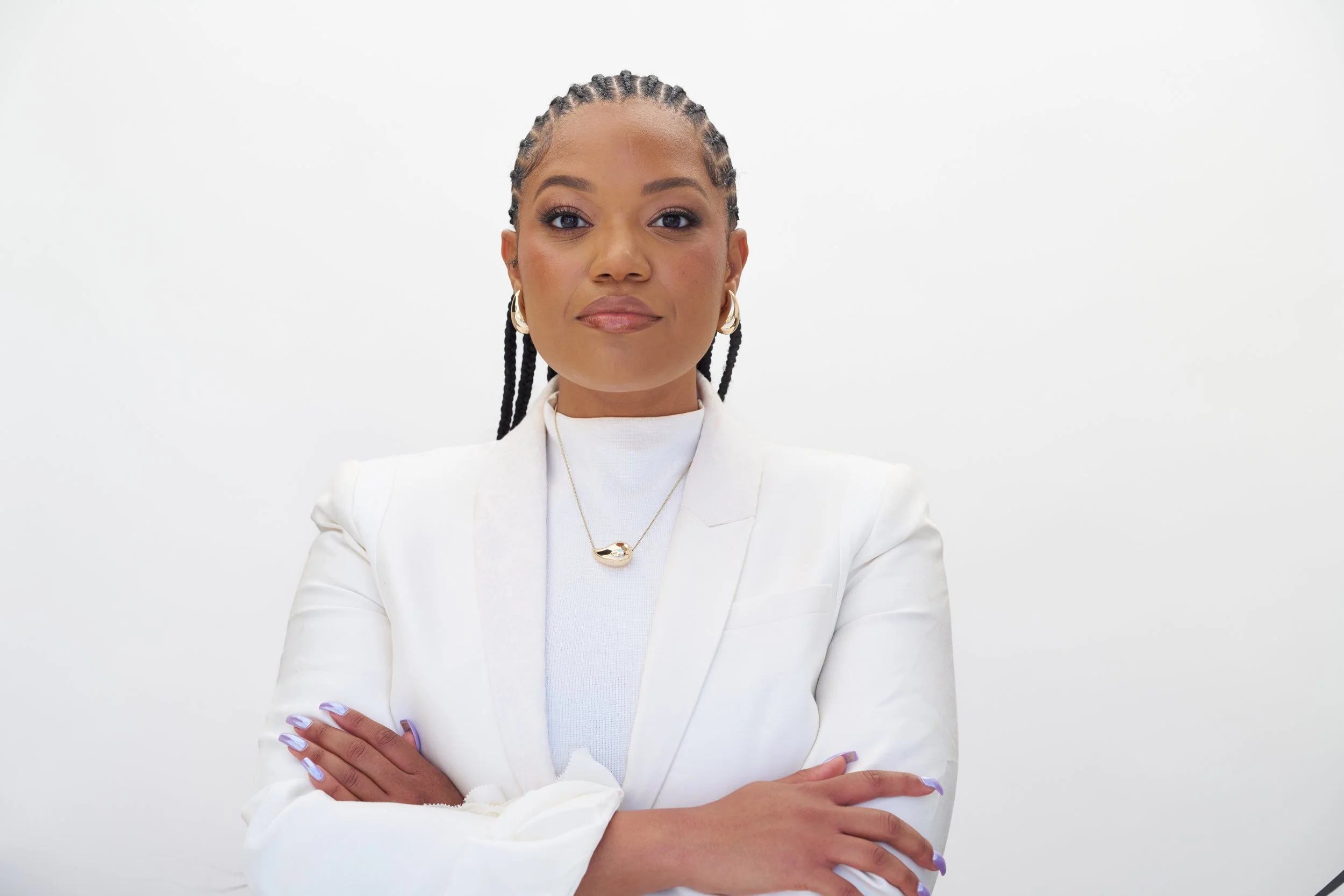 A confident woman with long braids, wearing a white blazer and jewelry, standing with crossed arms against a plain white background.