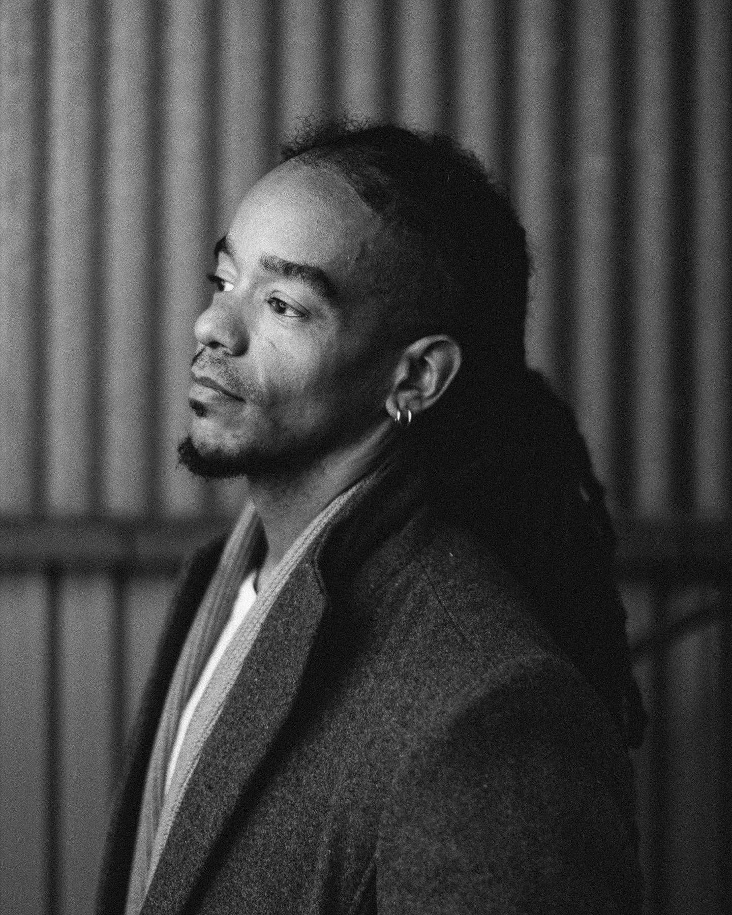 Black and white portrait of a man with a goatee and earrings, looking to the side, with a background of vertical wooden slats.