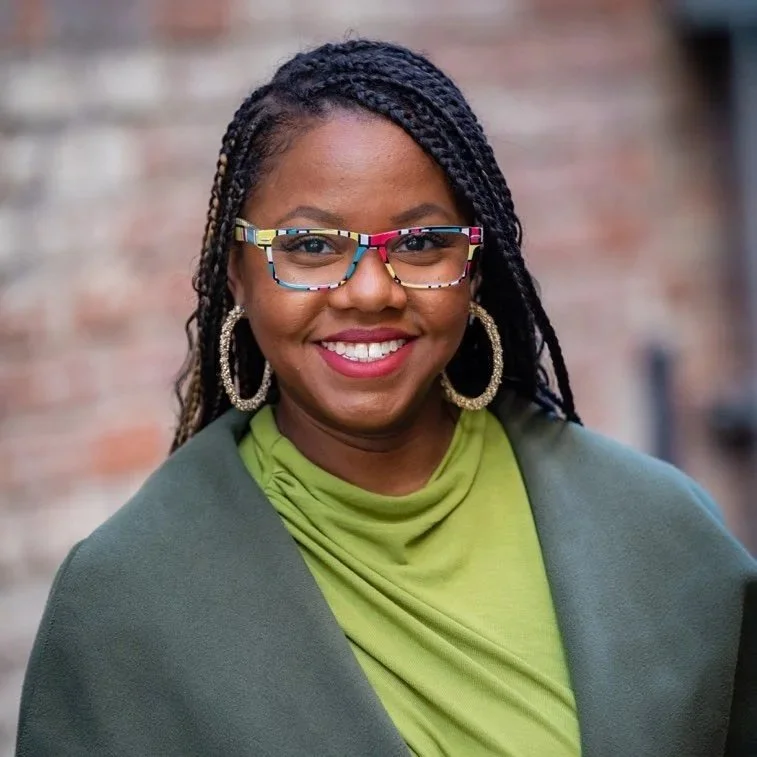 Smiling woman with braided hair, colorful glasses, hoop earrings, wearing a green top and a gray shawl, standing outdoors with a brick wall background.