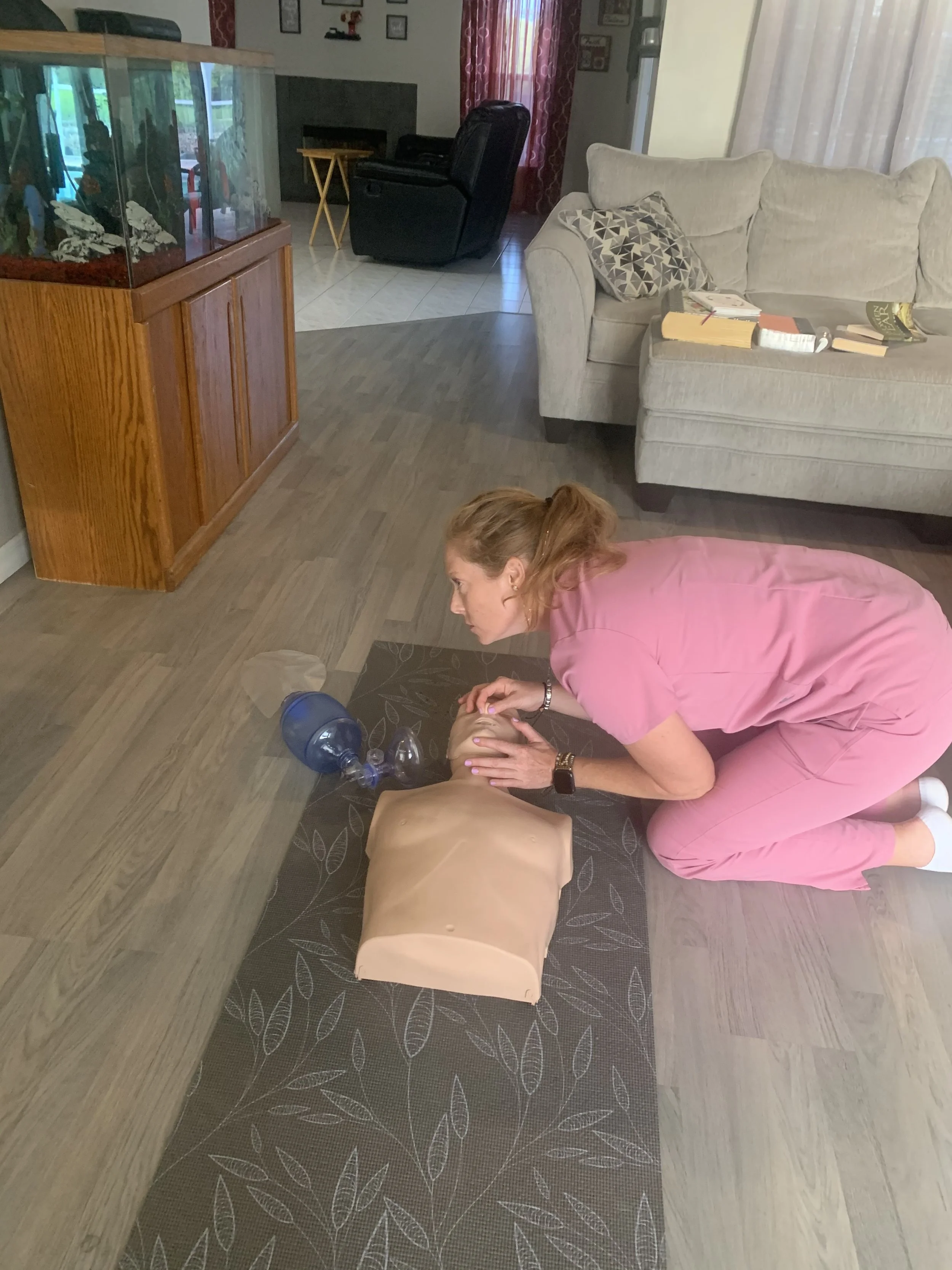 Woman in pink scrubs practicing CPR on a mannequin in a living room.