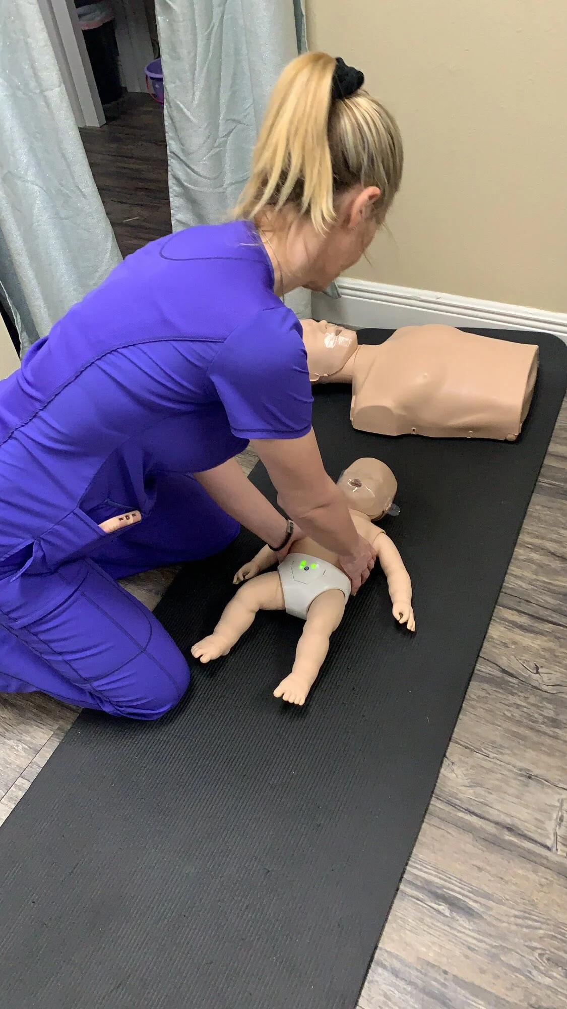 A woman in purple medical scrubs performing CPR on a baby mannequin on a black mat, with a larger mannequin on the table nearby.