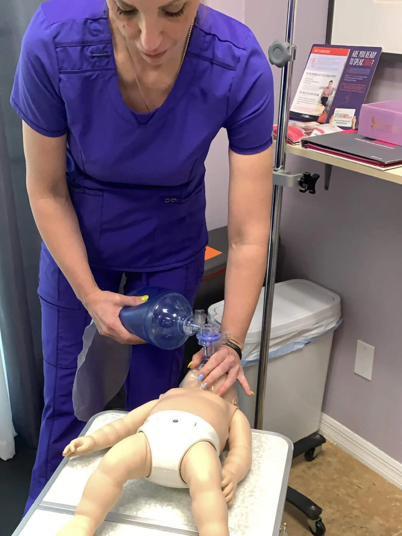 A medical professional in purple scrubs performing CPR on a baby mannequin using a bag valve mask to provide ventilation.