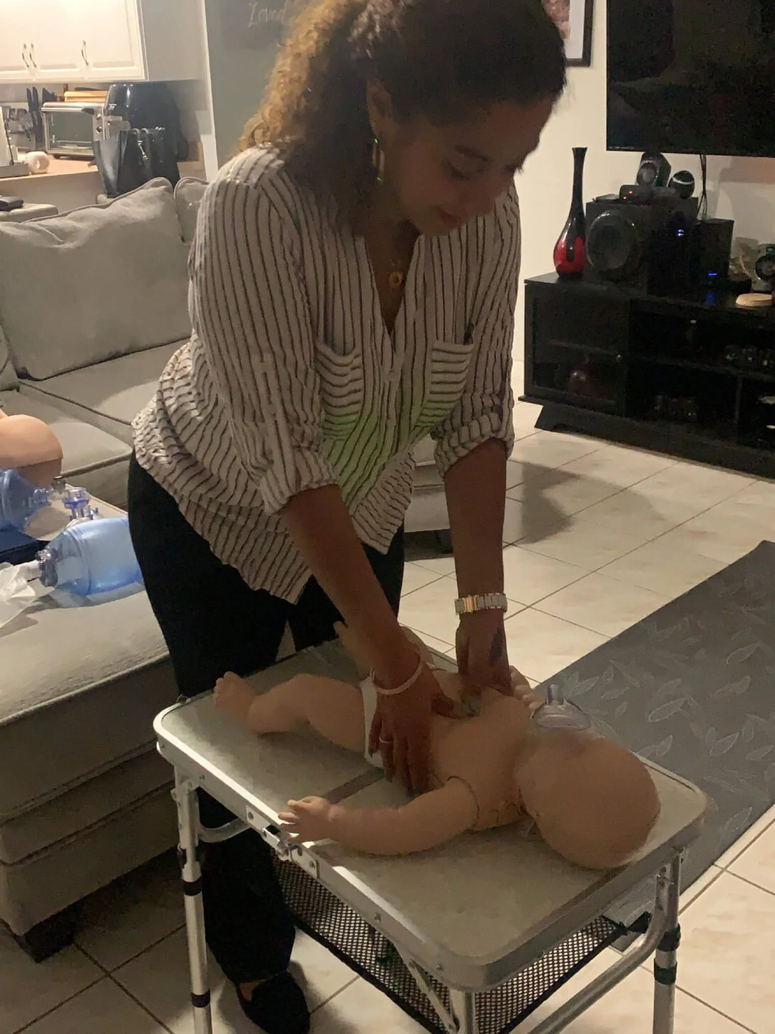 A woman practicing infant CPR on a baby doll on a table in a living room.