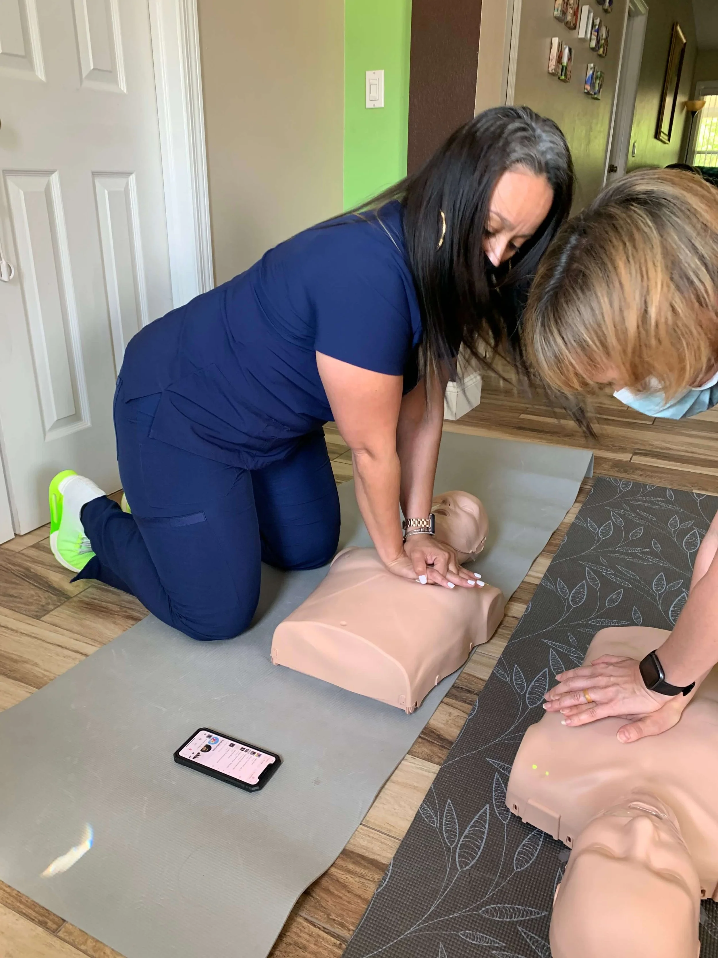 Two women practicing CPR on mannequins inside a home, with a smartphone nearby showing instructions.