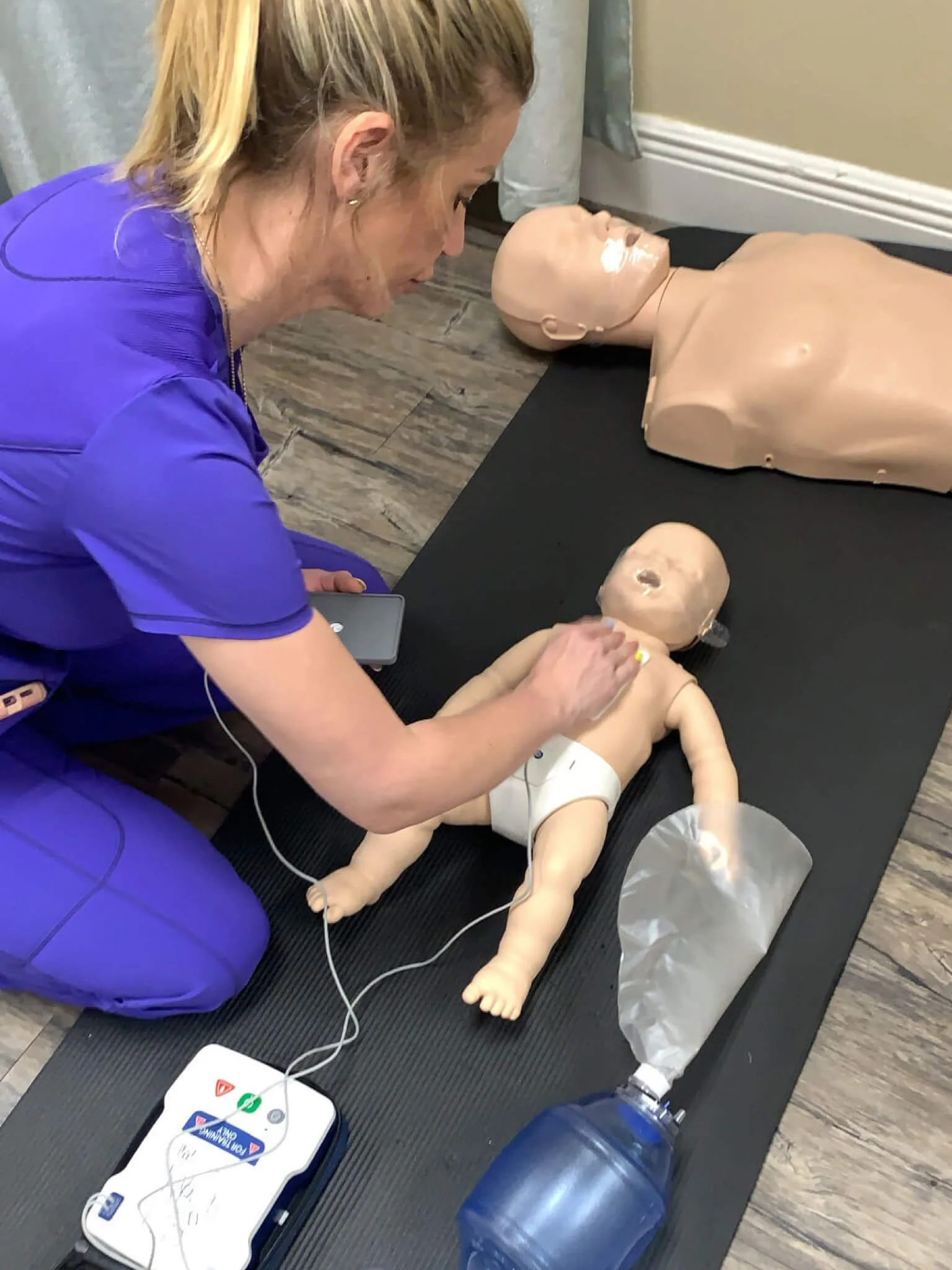 Woman in purple scrubs practicing CPR on a baby medical training doll with adult and infant mannequins nearby.
